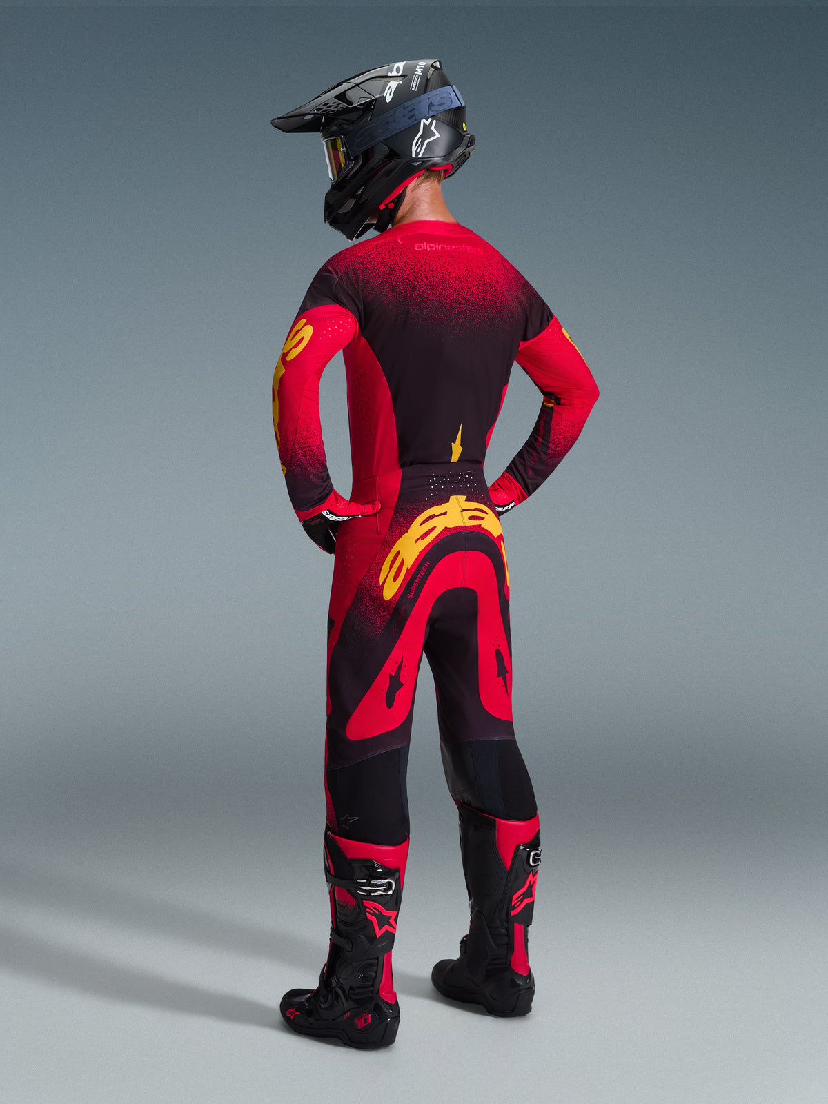 Motocross-Fahrer von hinten in Alpinestars 2026 Supertech Scenz Hose in Rot Schwarz Gelb, mit einem großen gelben Logo auf dem Gesäß, kombiniert mit einem passenden rot-schwarzen Jersey mit Farbverlauf, schwarzem Helm und roten Motocross-Stiefeln, professionelles Rennausrüstungs-Set für Offroad-Performance.