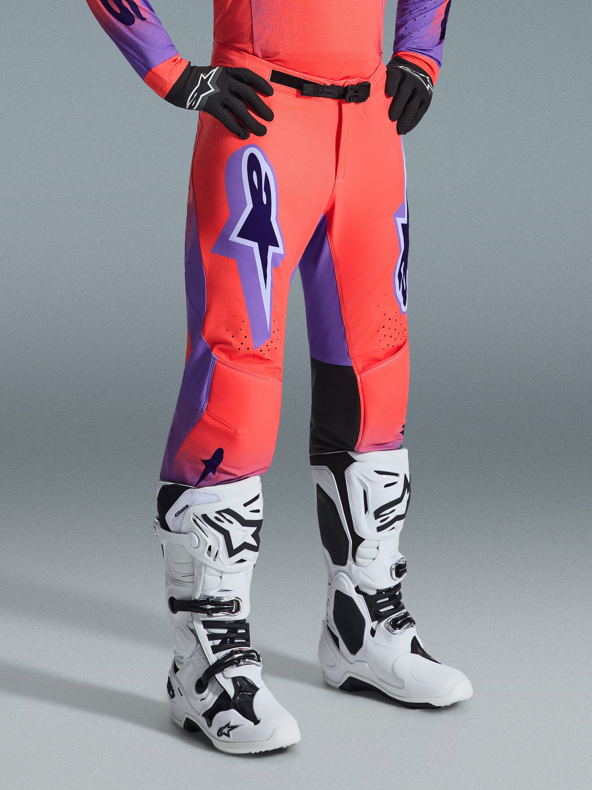 Alpinestars 2026 Supertech Scenz Hose, Motocross-Hose, Hot Coral und Lila, kombiniert mit weiß-schwarzen Motocross-Stiefeln und schwarzen Handschuhen, großes lila Alpinestars-Logo, schwarze Leder-Innenknie-Panels, athletisches Race-Fit-Design