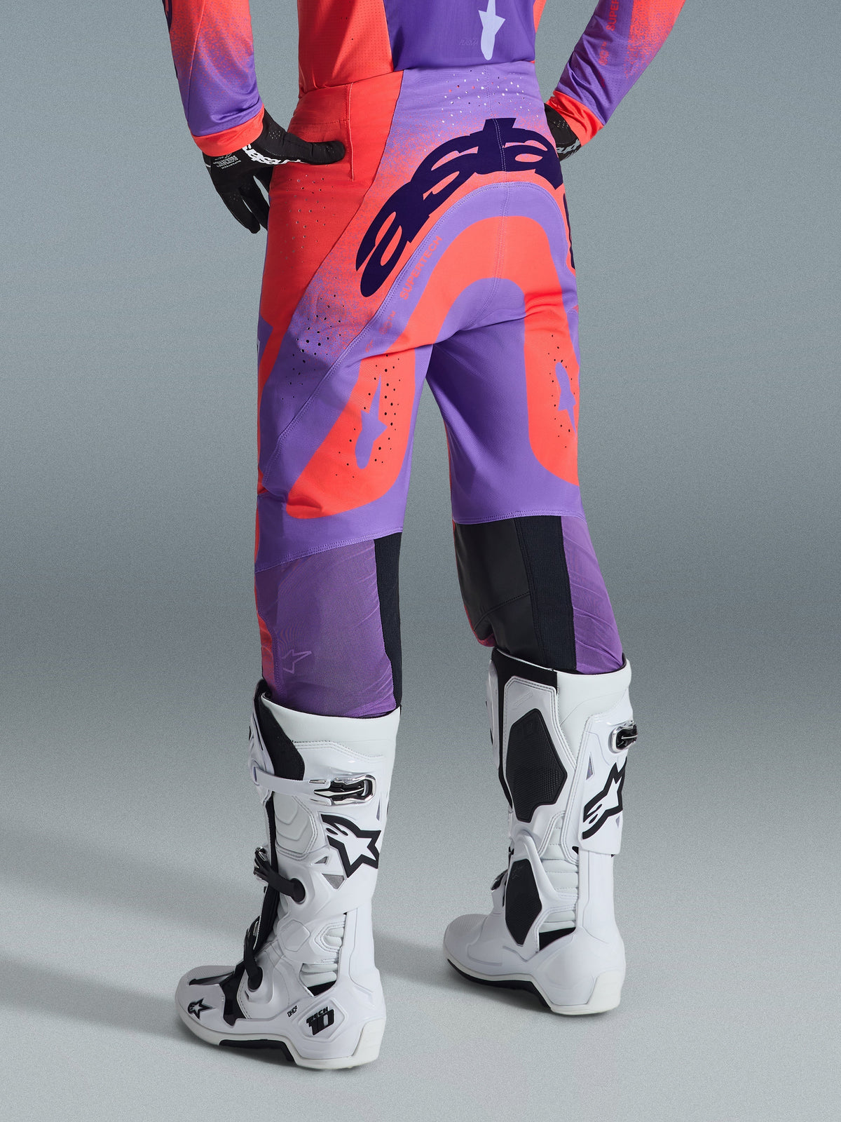 Alpinestars 2026 Supertech Scenz Hose, Motocross-Hose, Hot Coral Purple, Rückansicht mit markantem Branding am Gesäß, leuchtende korallenrote und lilafarbene Panels mit perforierten Zonen für die Luftzirkulation, getragen mit weiß-schwarzen Rennstiefeln