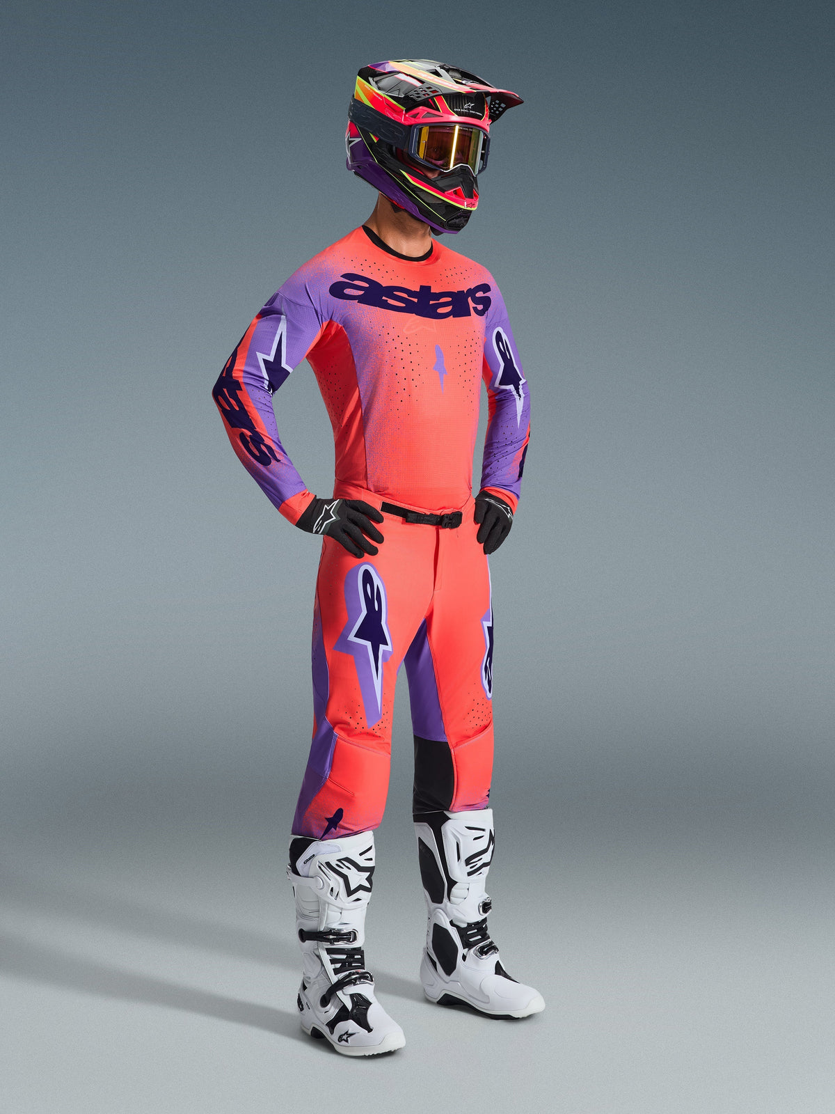 Motocross-Fahrer in kompletter Rennausrüstung, Alpinestars 2026 Supertech Scenz Hose in Hot Coral Purple, passendes Trikot in Koralle und Lila, schwarz-pinker Helm, weiß-schwarze Stiefel, professionelles Offroad-Rennset.