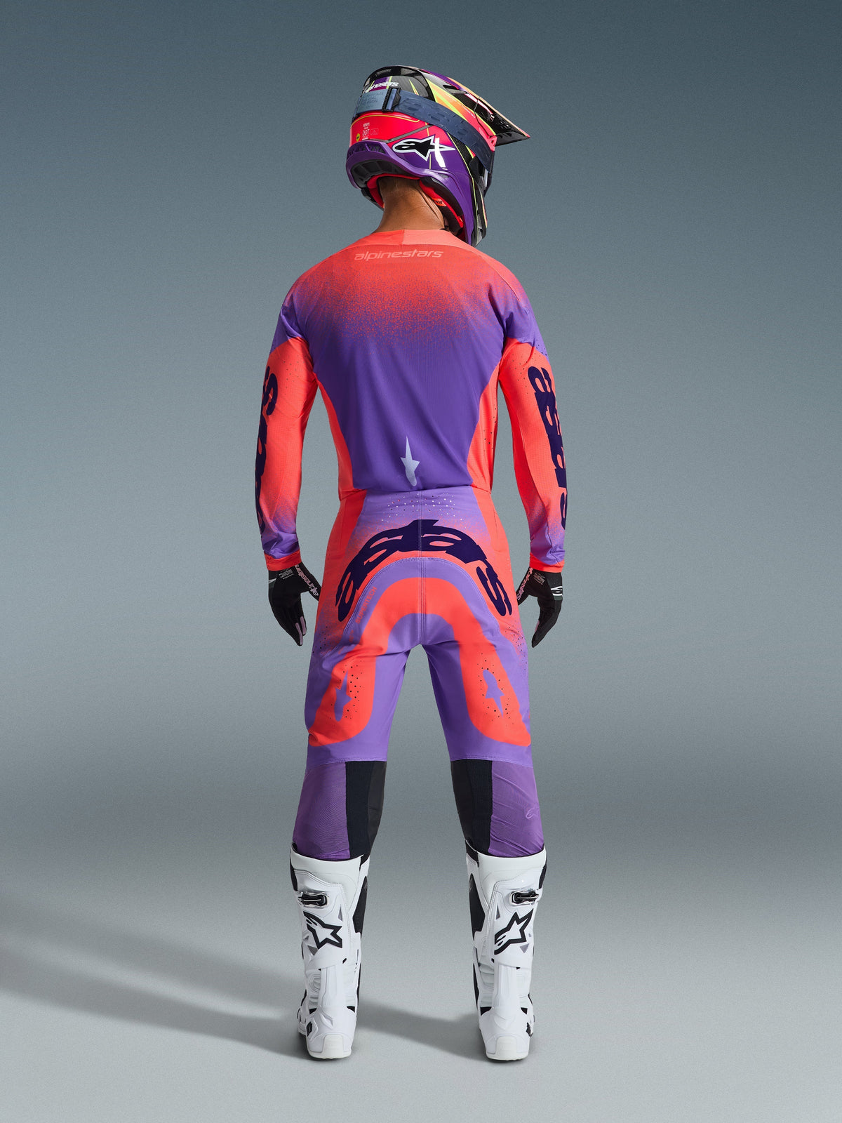 Alpinestars 2026 Supertech Scenz Hose, Motocross-Hose, Hot Coral und Lila, Rückansicht eines Motocross-Fahrers in kompletter Rennausrüstung, mit lebhaftem Farbverlauf-Design und markanten Logos auf der Rückseite, kombiniert mit passendem Jersey, Helm und weißen Stiefeln.