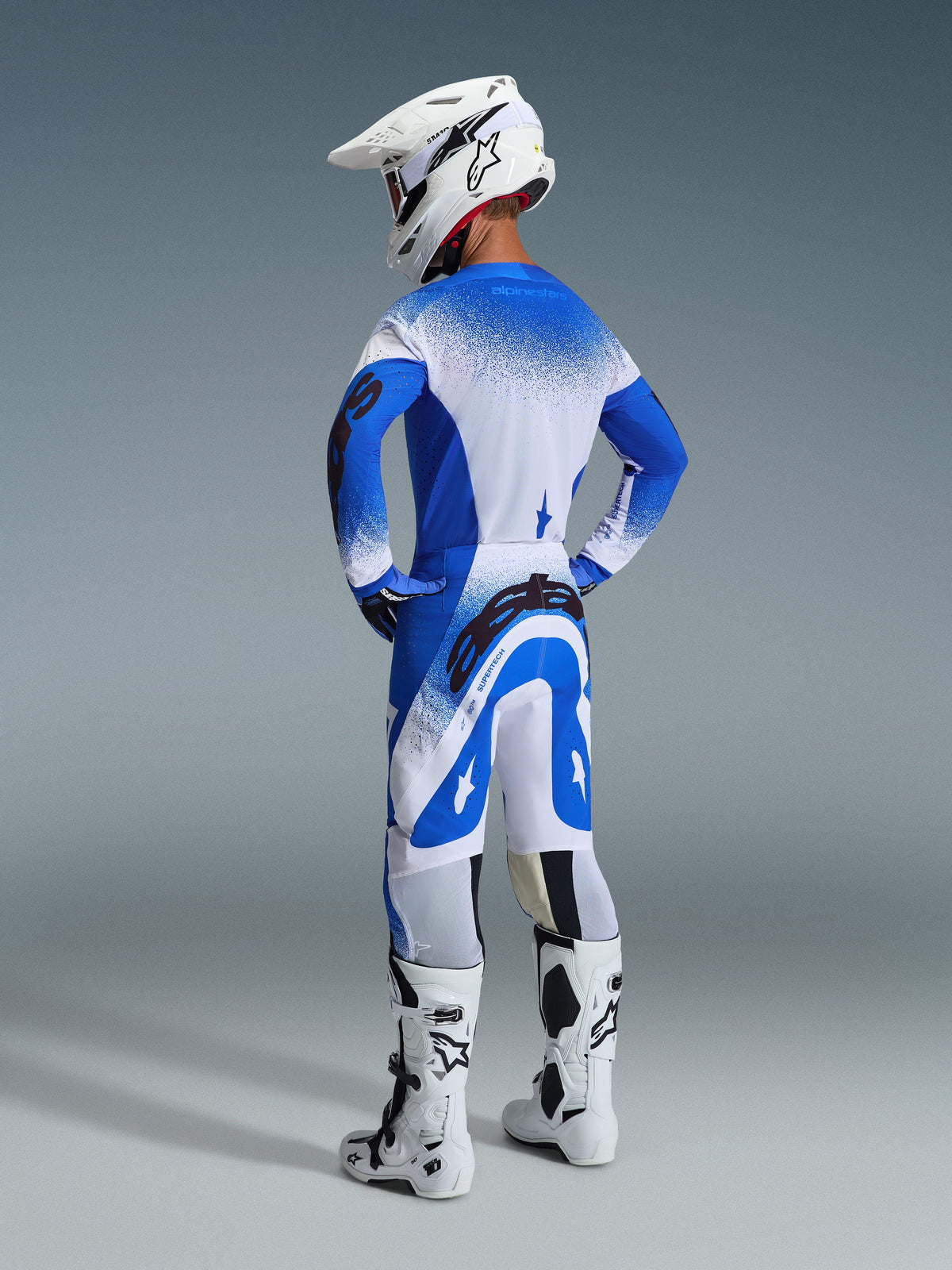 Motocross-Fahrer in der Rückansicht mit der 2026 Supertech Scenz Hose und dem Trikot in Ucla Blau-Weiß, weißer Helm mit Alpinestars-Logo, weiß-schwarze Motocross-Stiefel, professionelle Offroad-Rennausrüstung.