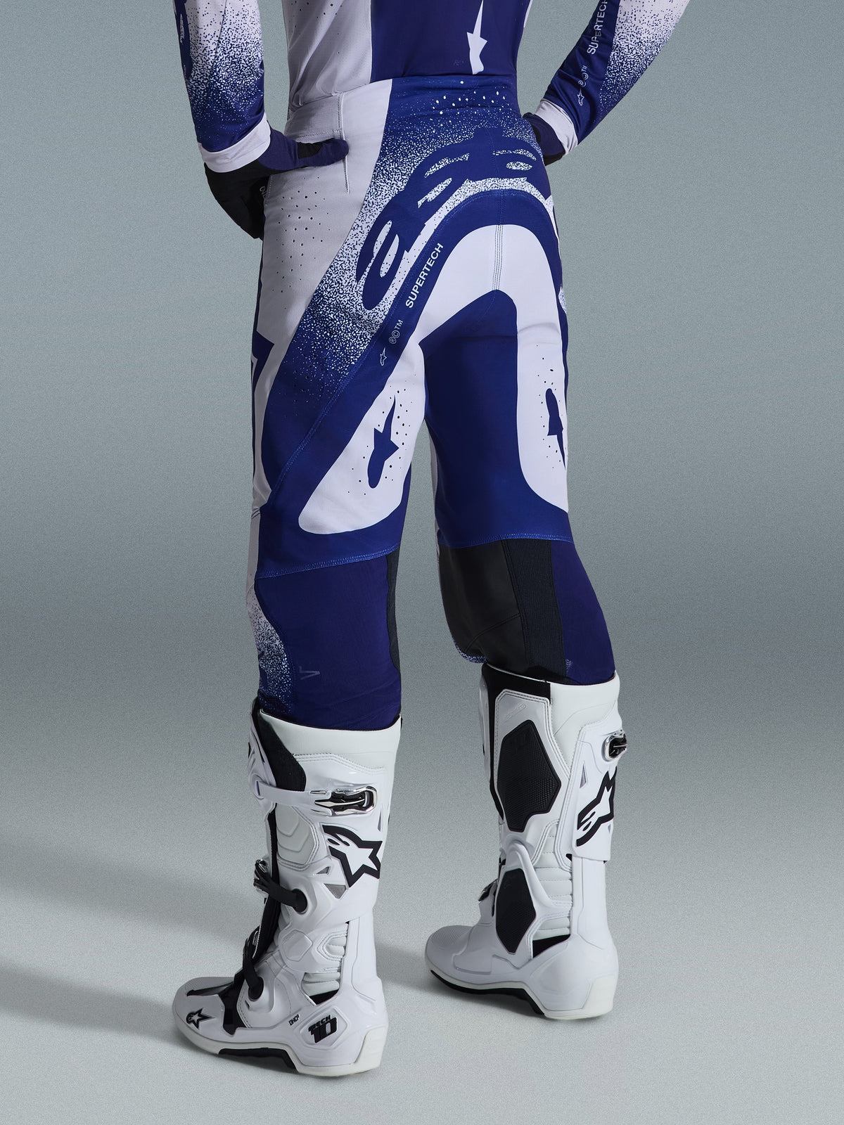 Alpinestars 2026 Supertech Scenz Hose, Motocross-Hose, Hellgrau und Dunkelblau, Rückansicht getragen mit passendem Trikot und weißen Stiefeln, markante blaue Grafiken mit gesprenkelten Akzenten, athletische Performance-Passform für den Offroad-Rennsport.
