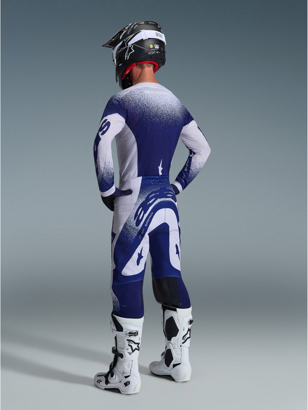 Alpinestars 2026 Supertech Scenz Hose, Motocross-Hose, Hellgrau und Dunkelblau, Rückansicht getragen mit passendem Trikot und weißen Stiefeln, mit Punkt-Grafiken mit Farbverlauf und athletischer Performance-Passform für den Offroad-Rennsport.