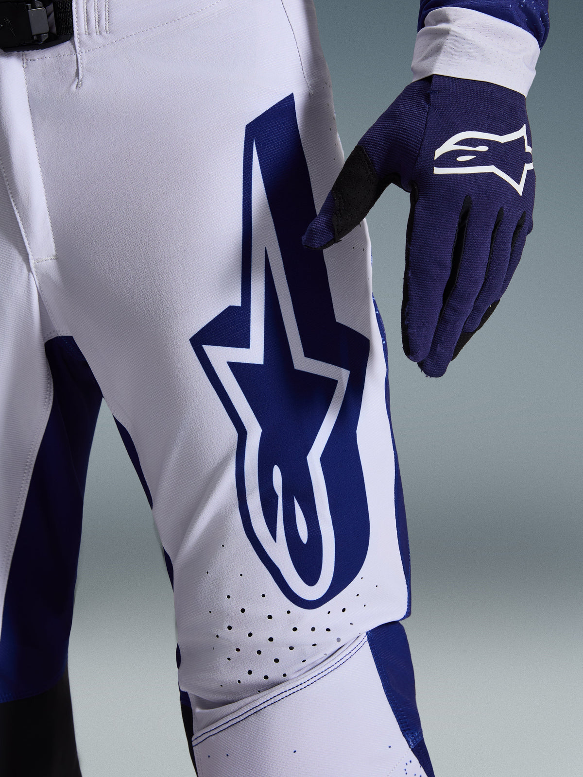 Alpinestars 2026 Supertech Scenz Hose, Motocross-Hose, Hellgrau und Dunkelblau, Nahaufnahme des Oberschenkels mit großem dunkelblauem Alpinestars-Logo, perforierte Belüftungszonen und Stretch-Ripstop-Gewebe, getragen mit passendem dunkelblauem Motocross-Handschuh