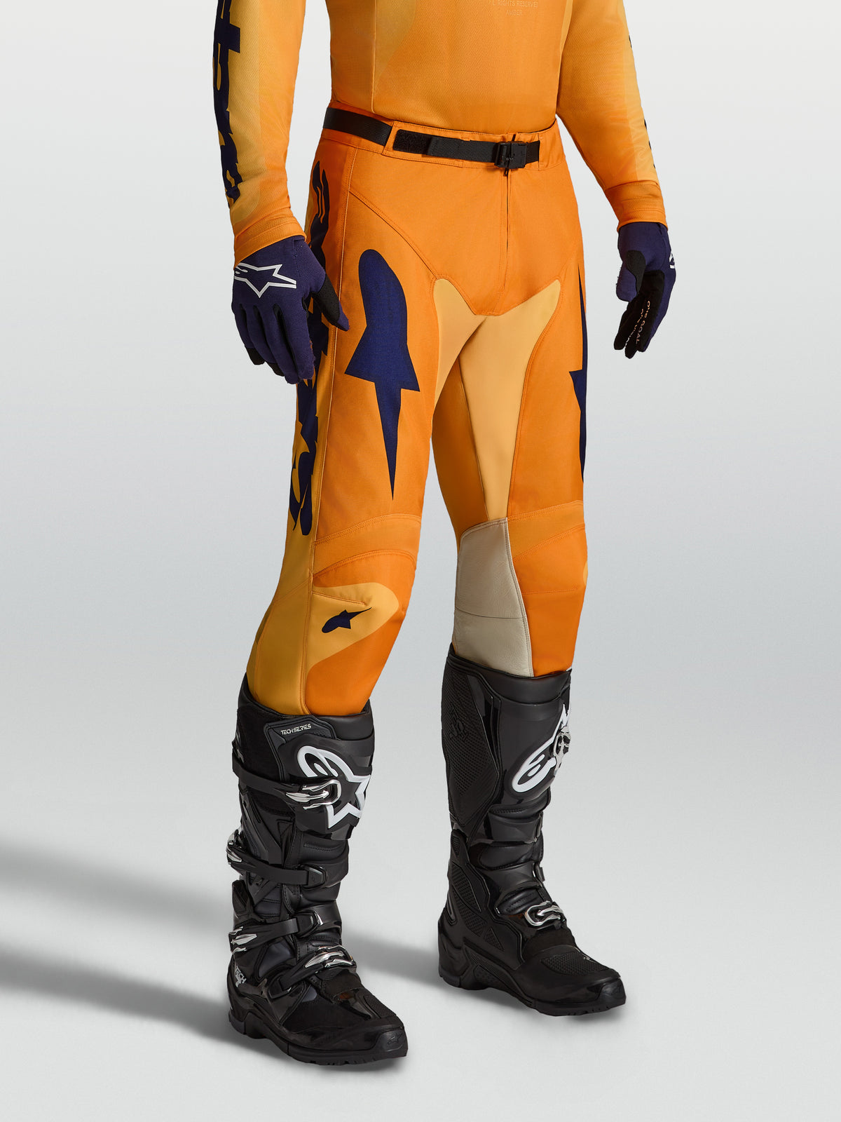 Alpinestars Racer Amber Hose, Motocross-Hose für Herren, Orange und Dunkelblau, Stretch-Einsätze und vorgekrümmte Beinform, getragen mit schwarzen Stiefeln.