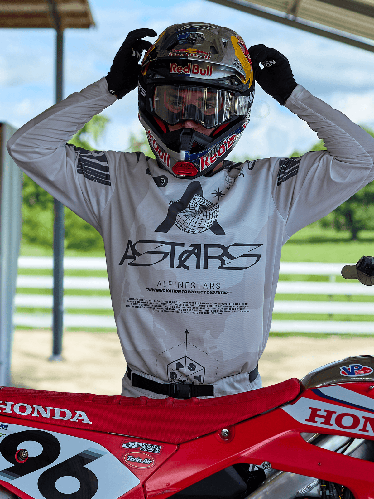 Motocross-Fahrer in Alpinestars 2025 Racer Hollow Ausrüstung, helles Camo-Trikot mit markantem ASTARS-Branding, justiert einen Red Bull Helm mit klarer Brille, neben einem roten Honda-Dirtbike in einer Offroad-Umgebung.