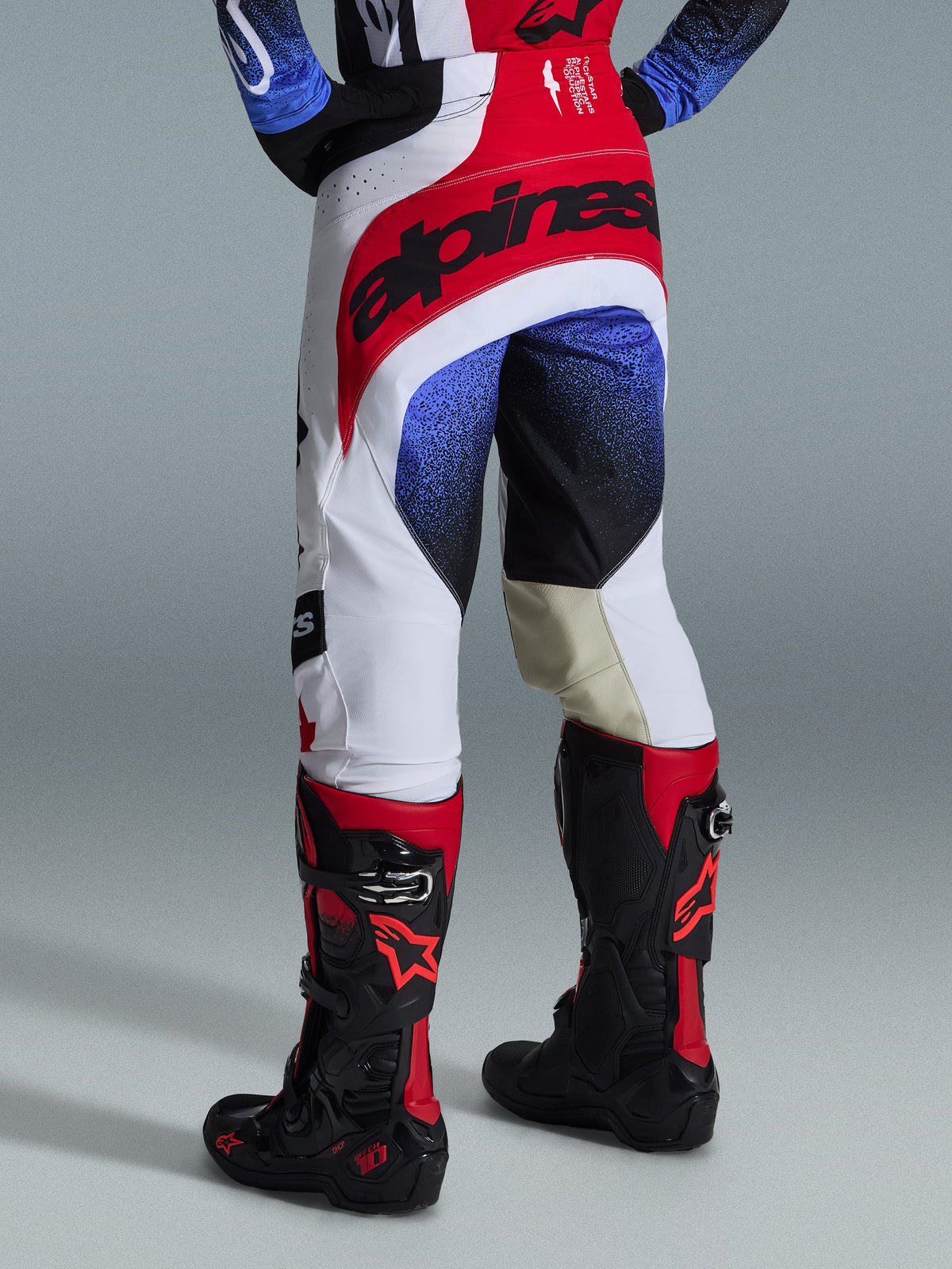 Alpinestars 2026 Techstar Knif Pants, Motocross Pants, Black White and Red with blue accents, rear view showing bold branding on the waist and tan leather inner knee panels, worn with black and red boots, high-performance racing gear for off-road competition