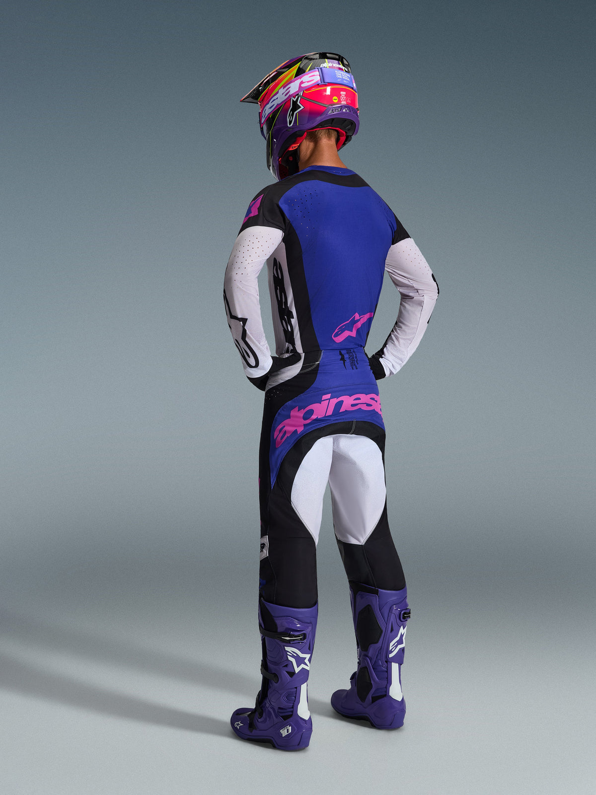 Motocross-Fahrer in der Rückansicht, trägt das Alpinestars 2026 Techstar Knif Gear Set, blaues Jersey mit weißen perforierten Ärmeln und pinkfarbenen Logos, schwarz-weiße Hose mit pinkfarbenem Branding auf der Rückseite, kombiniert mit lila Stiefeln und einem leuchtend lila-pinkfarbenen Helm, professionelles Motocross-Rennoutfit.