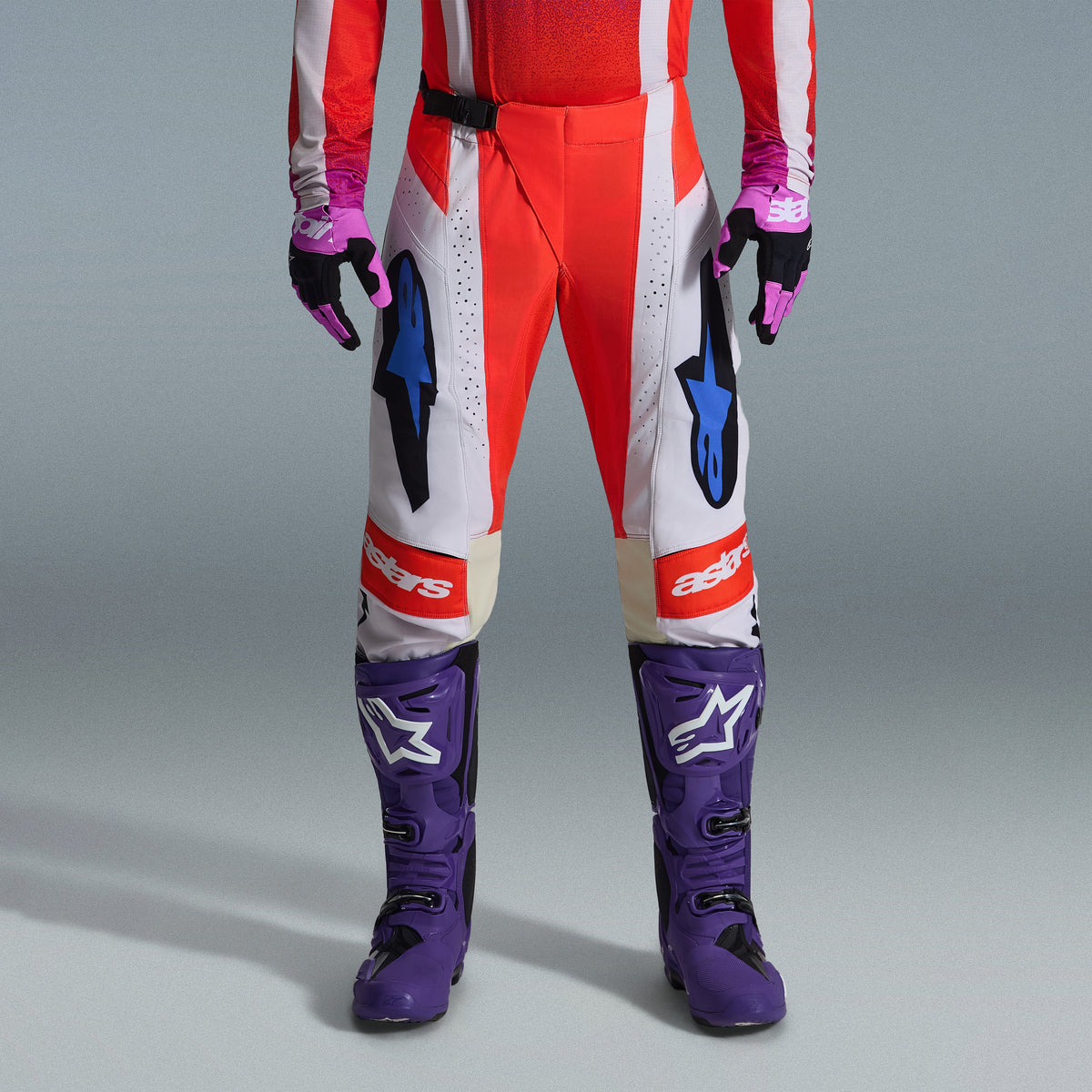 Alpinestars 2026 Techstar Knif Bekleidungsset, Unterkörper eines Motocross-Fahrers in rot-weißer Hose mit blauen Akzenten, leuchtend lila Rennstiefel mit weißen Logos und passenden schwarz-lila Handschuhen, professionelle Offroad-Rennausrüstung