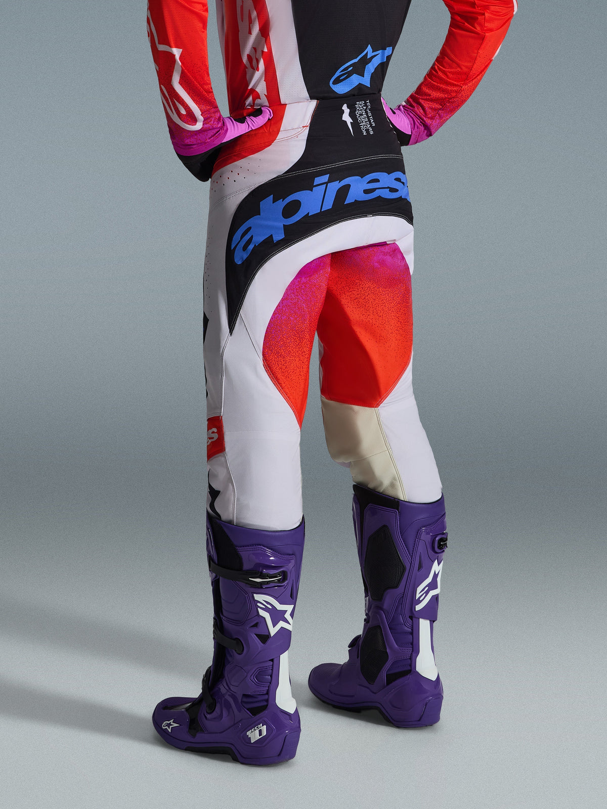 Alpinestars 2026 Techstar Knif Hose und Ausrüstungsset, Rückansicht eines Motocross-Fahrers mit weißer Hose mit schwarzen Einsätzen, pinken und roten Farbverlaufsakzenten und einem großen blauen Alpinestars-Logo auf der Sitzfläche, kombiniert mit lila Tech 10 Stiefeln und einem passenden rot-schwarzen Jersey.