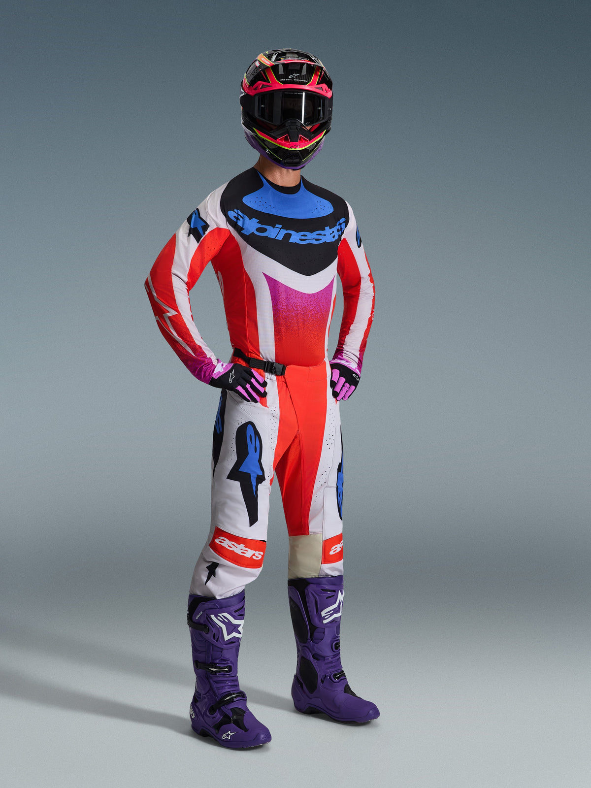 Motocross-Fahrer in kompletter Alpinestars Rennausrüstung, 2026 Techstar Knif Trikot in Weiß und Rot mit blauen und pinken Akzenten, passende Motocross-Hose, lila Stiefel und ein mehrfarbiger Helm, koordiniertes professionelles Outfit für Offroad-Performance