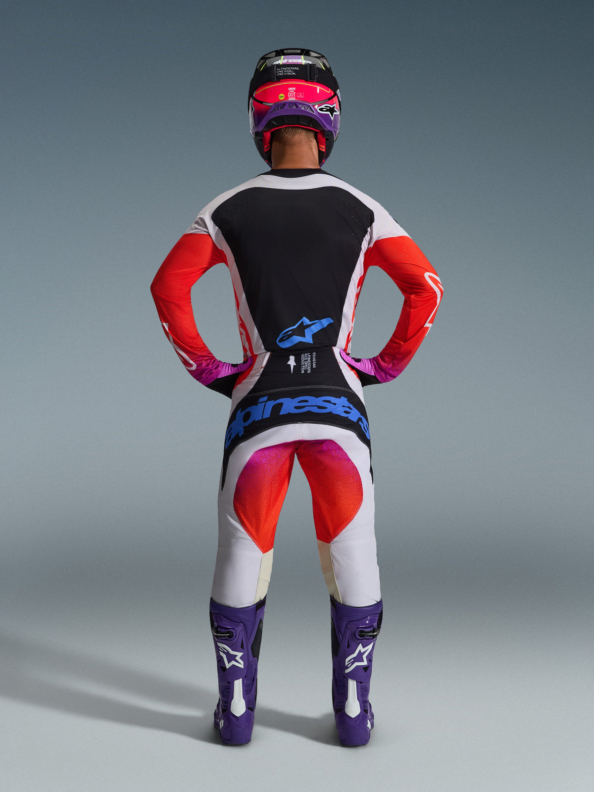 Motocross-Fahrer in kompletter 2026 Techstar Knif Rennausrüstung von hinten, hellgraues Trikot mit schwarzem Rückeneinsatz und orangefarbenen Ärmeln, passende weiß-orangefarbene Hose mit blauem Alpinestars-Branding, lila Stiefel und schwarz-pinker Helm, abgestimmtes professionelles Outfit für den Offroad-Rennsport