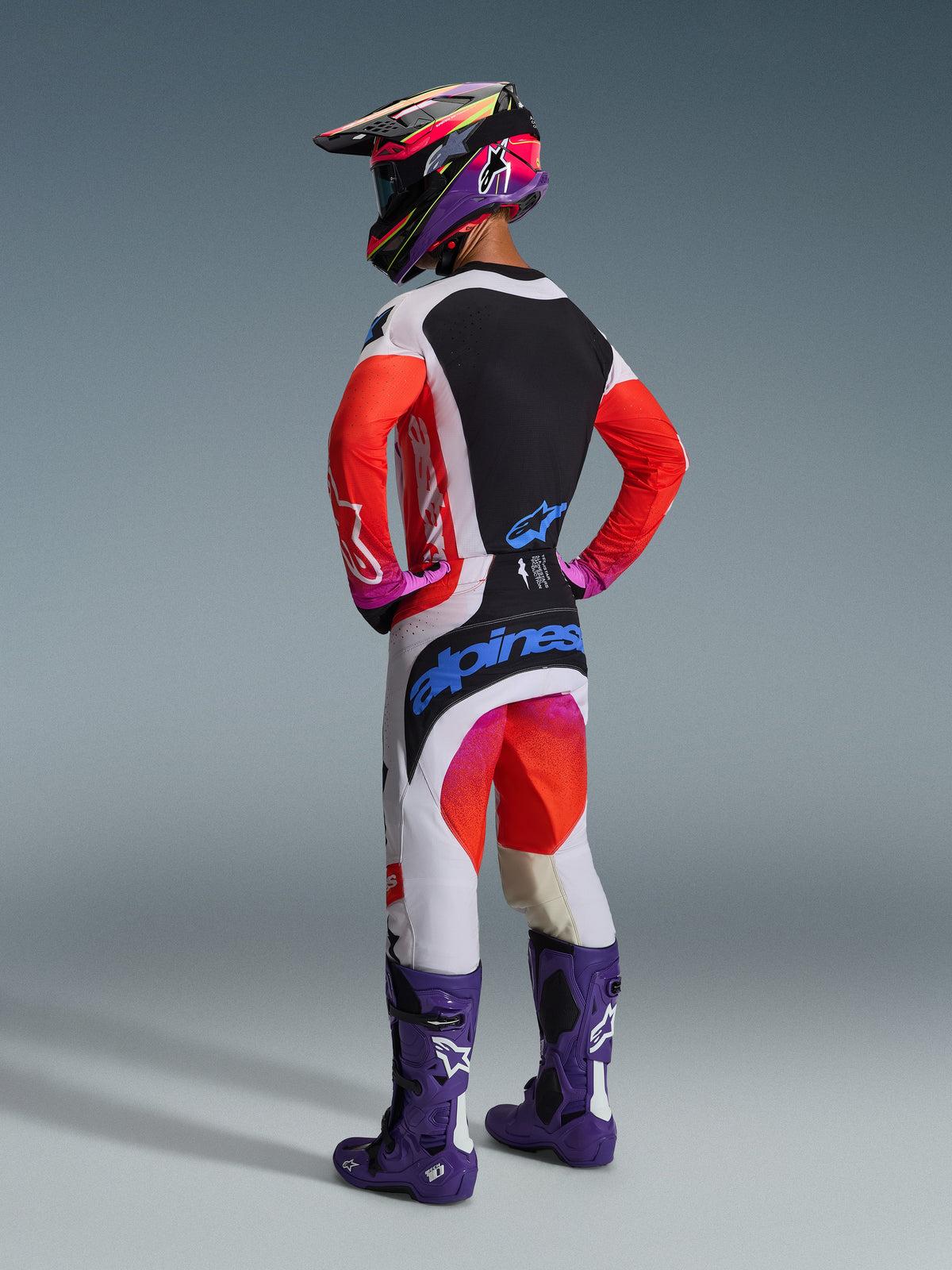 Motocross-Fahrer in der Rückansicht mit dem Alpinestars 2026 Techstar Knif Gear Set, weißes Jersey mit roten Ärmeln und schwarzem Panel, weiße Hose mit roten und lila Akzenten und blauem Alpinestars-Branding, lila Rennstiefel und passender Helm, professionelle Offroad-Rennausrüstung