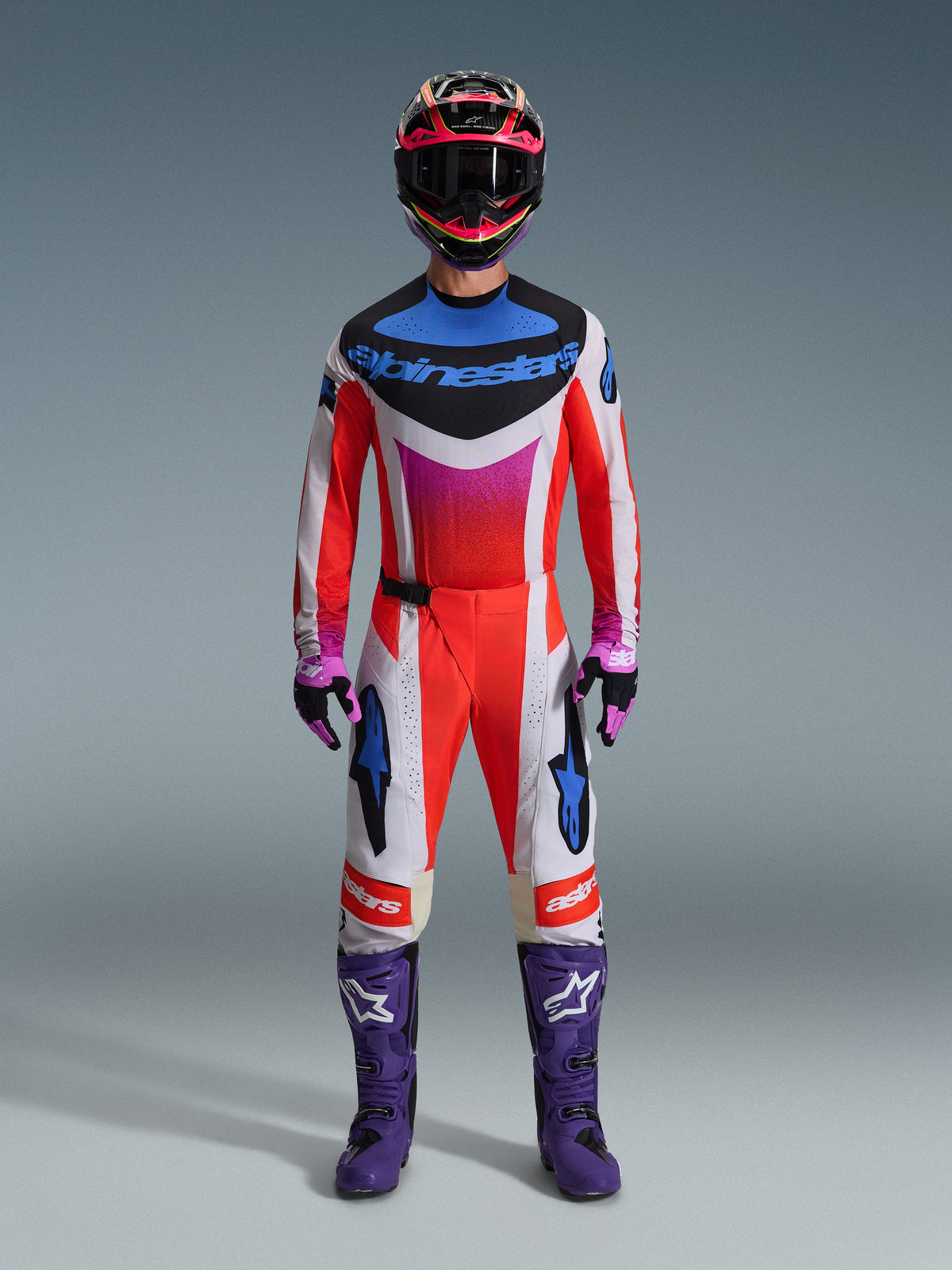 Motocross-Fahrer in kompletter Rennausrüstung, Alpinestars 2026 Techstar Knif Trikot und passende Hose in Weiß und Orange mit blauen und lila Akzenten, lila Rennstiefel mit weißem Logo und ein mehrfarbiger Helm, aufeinander abgestimmtes Outfit für Offroad-Rennsportwettbewerbe