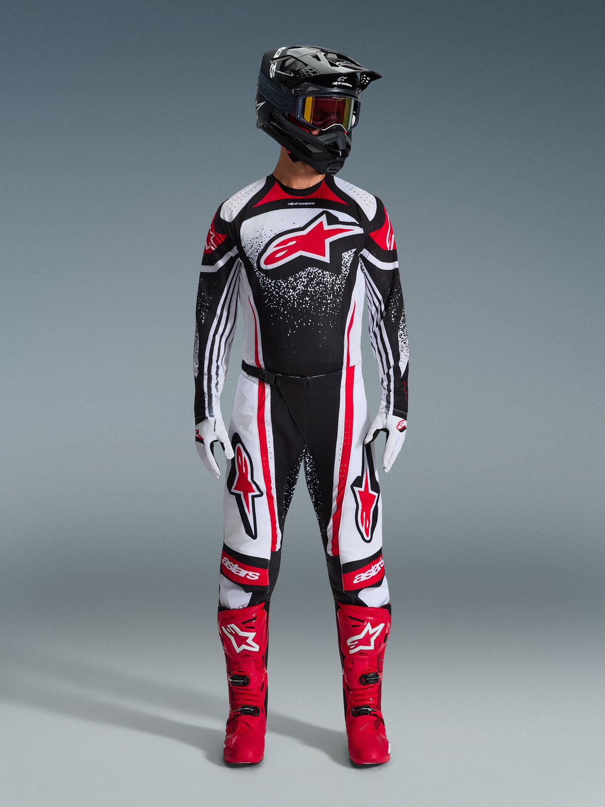 Alpinestars 2026 Techstar Nomur Gear Set, Motocross-Fahrer in kompletter Rennausrüstung, Design in Schwarz, Weiß und Rot mit weißem Sprenkelmuster, Jersey mit rotem Astars-Logo, dazu passende weiß-rote Hose, leuchtend rote Stiefel und schwarzer Helm mit Schutzbrille, abgestimmte High-Performance-Motocross-Ausrüstung