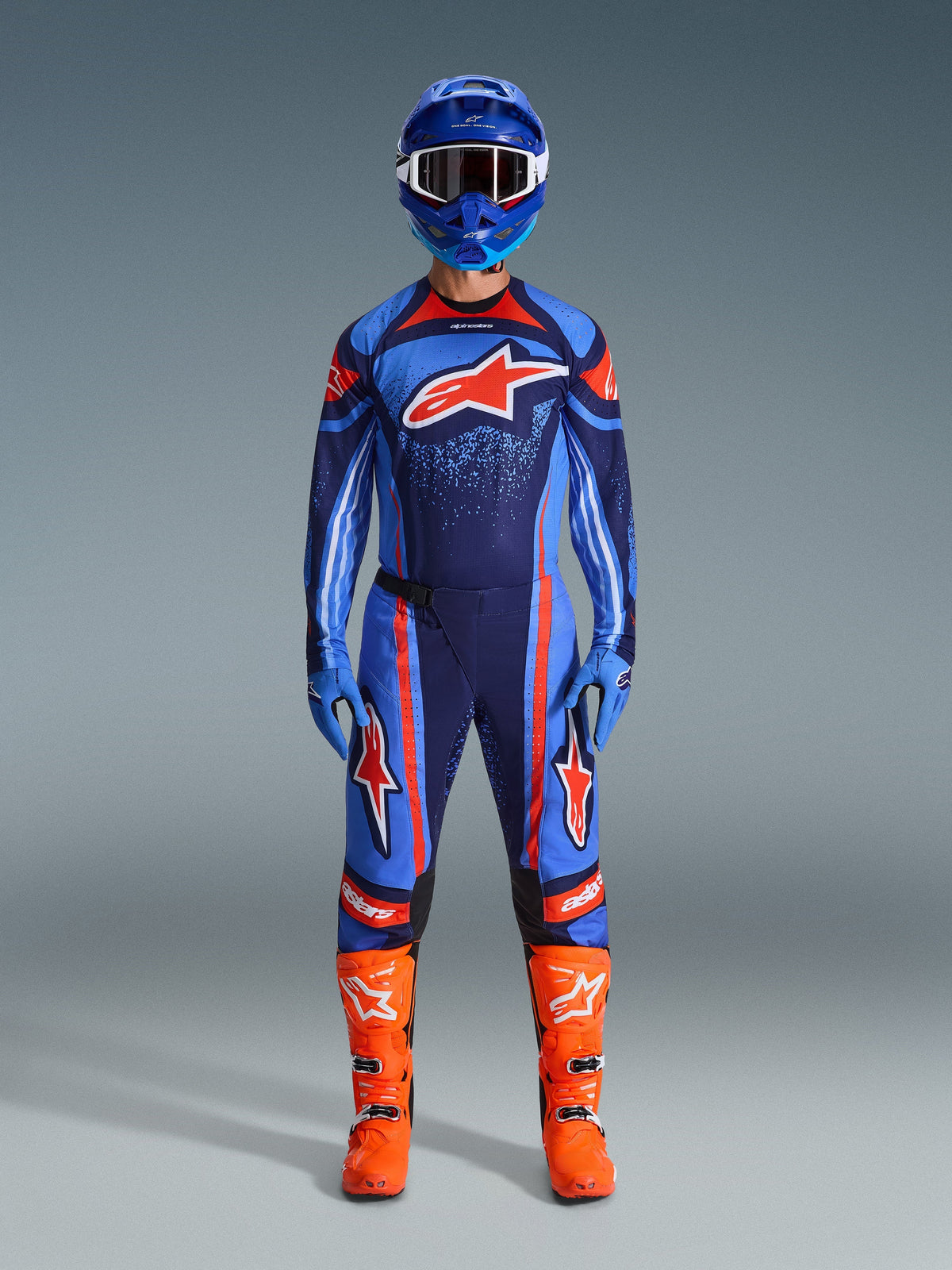 Alpinestars 2026 Techstar Nomur gear set, Motocross rider in full racing kit, Dark Navy with Orange and UCLA Blue accents, matching jersey and pants with large logos, blue helmet and bright orange boots, coordinated off-road competition gear.