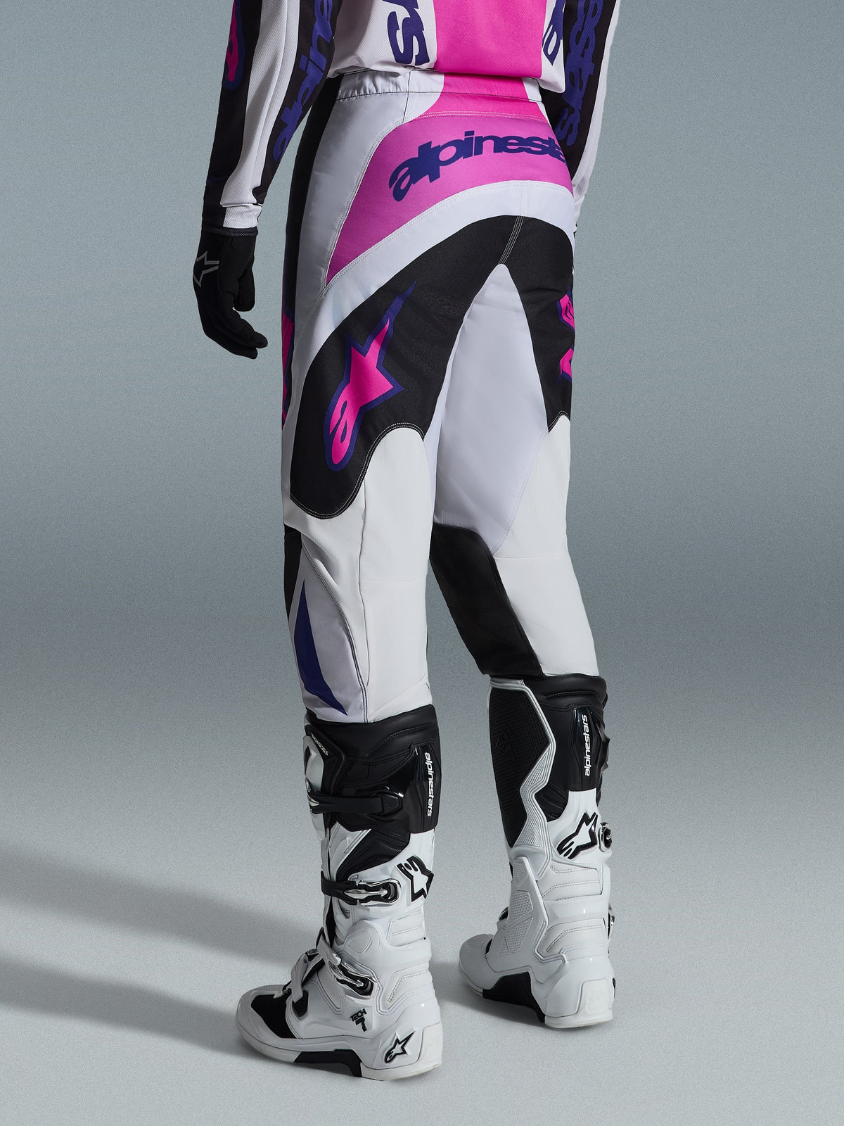 Alpinestars 2026 Fluid Grid Hose, Motocross-Hose, Schwarz, Hellgrau und Lila, Rückansicht eines Fahrers mit weiß-schwarzen Stiefeln, pinke und lila Alpinestars-Logos, sportliche Passform für Offroad-Rennsport.