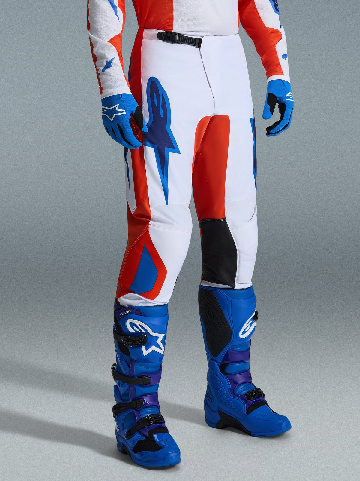 Alpinestars 2026 Fluid Grid Hose, Motocross-Hose, Orange und UCLA-Blau, kombiniert mit passenden blauen Handschuhen und Stiefeln, weiße Basis mit auffälligem Grafik-Branding, schwarze Knie-Innenpaneele und ergonomisches Design für den Offroad-Sport.
