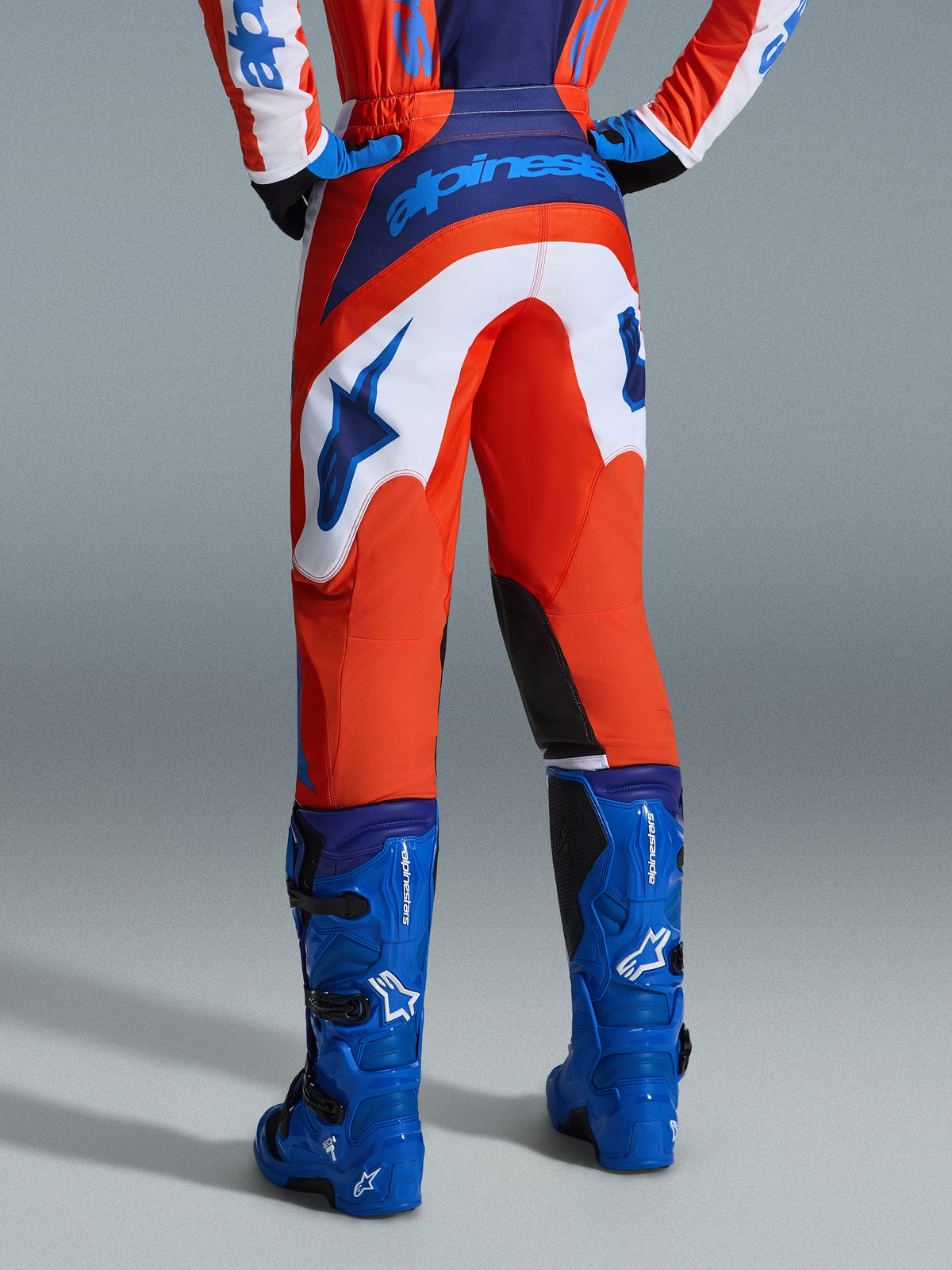 Alpinestars 2026 Fluid Grid Hose, Motocross-Hose, Orange und UCLA-Blau mit weißen Einsätzen, Rückansicht mit markantem blauem Branding auf der Gesäßpartie und Stern-Logo, kombiniert mit passenden blauen Motocross-Stiefeln, professionelle Offroad-Rennausrüstung