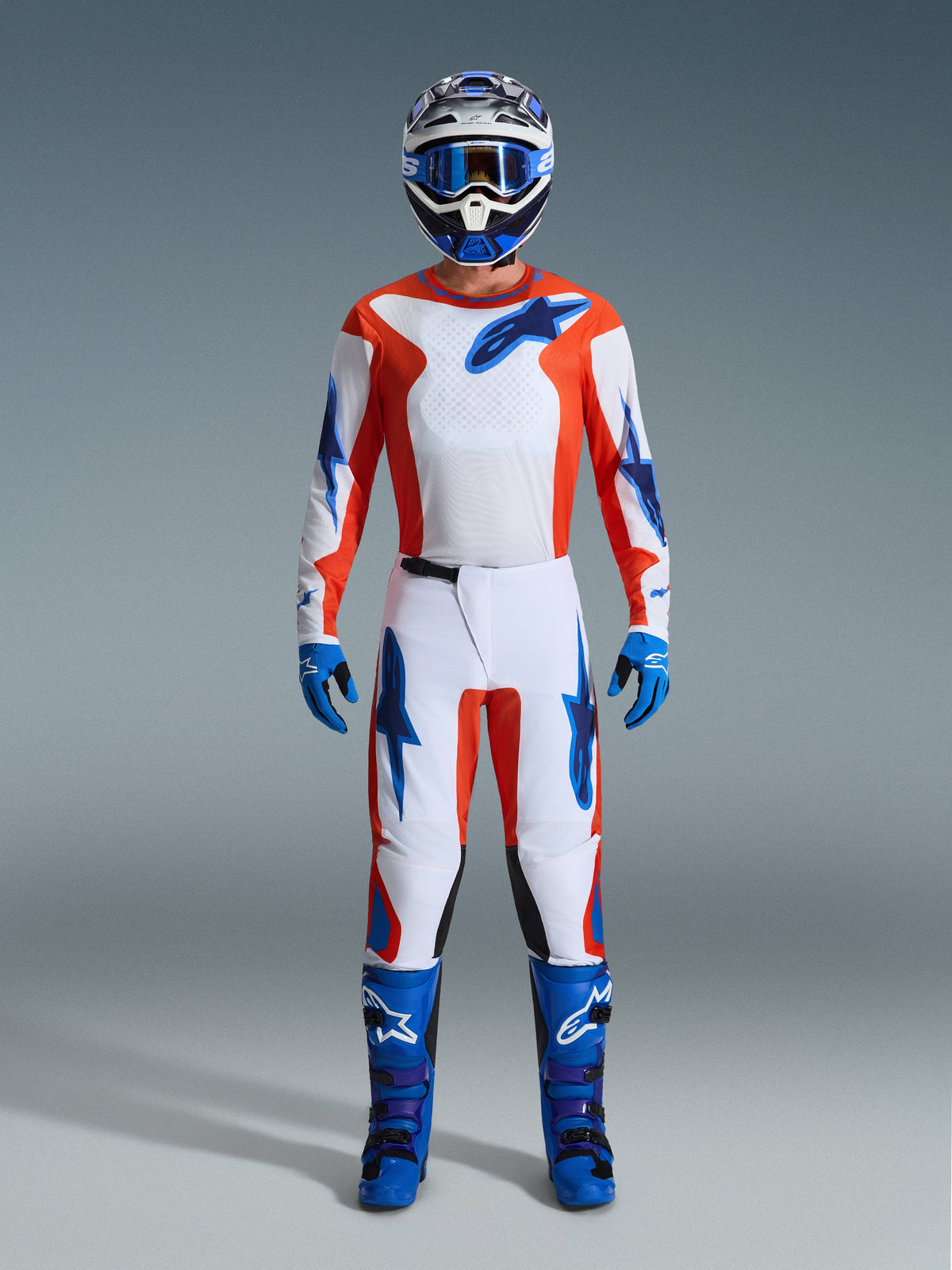 Motocross-Fahrer in kompletter Rennausrüstung, Alpinestars 2026 Fluid Grid Hose in Orange und UCLA Blau, weißes Trikot mit orangen Ärmeln und blauen Logos, passender blauer Helm und Brille, blaue Stiefel und blaue Handschuhe, abgestimmtes Outfit für Offroad-Rennsport