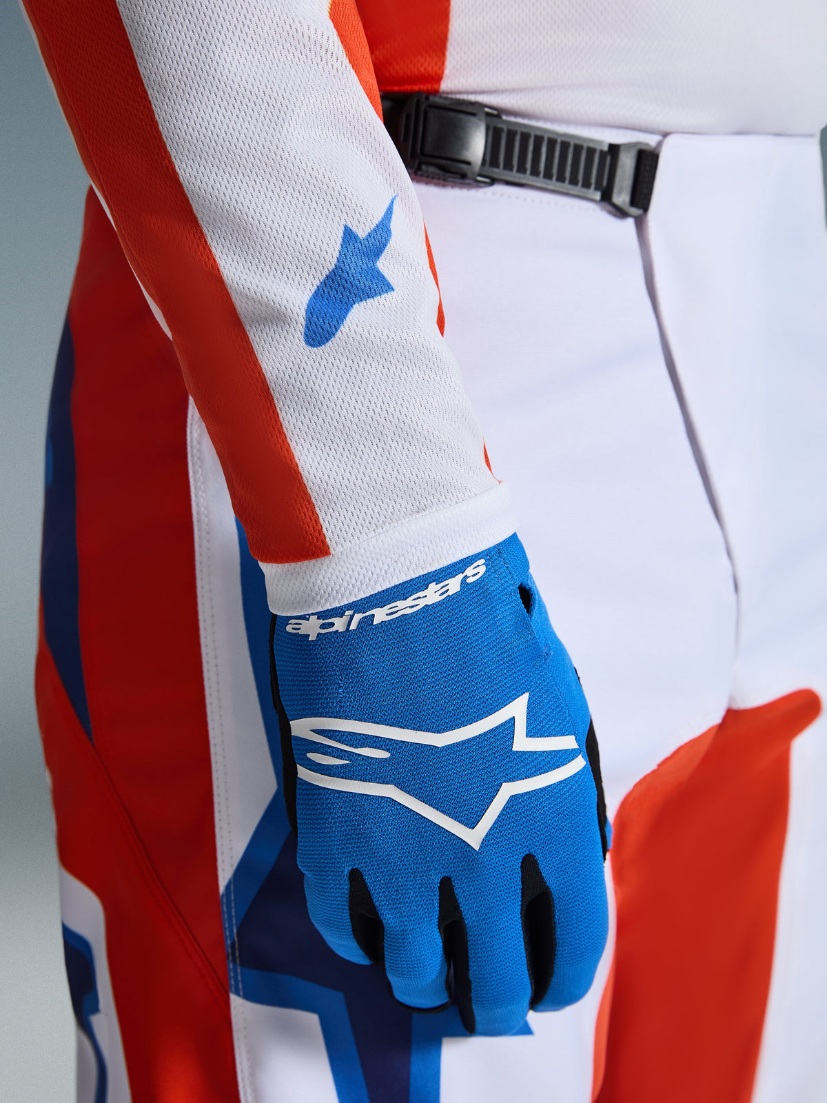 Alpinestars 2026 Fluid Grid Hose, Motocross-Hose, Orange und UCLA Blau, Nahaufnahme mit passendem blauem Handschuh und weißem Mesh-Trikotärmel mit blauen Logos, zeigt den Ratschenverschluss am Bund und die sportliche Passform.