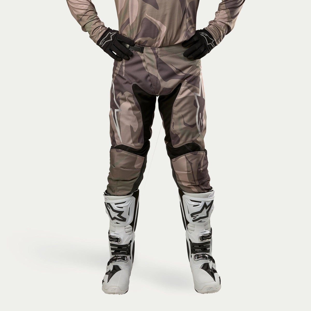 Alpinestars 2024 Racer Tactical Hose, Motocross-Hose, Military Green Camo Brown, mit großen weißen Alpinestars-Logos an den Oberschenkeln und schwarzen Innenbein-Einsätzen, getragen mit weiß-schwarzen Motocross-Stiefeln und schwarzen Handschuhen, passendes Camouflage-Trikot, Frontansicht.