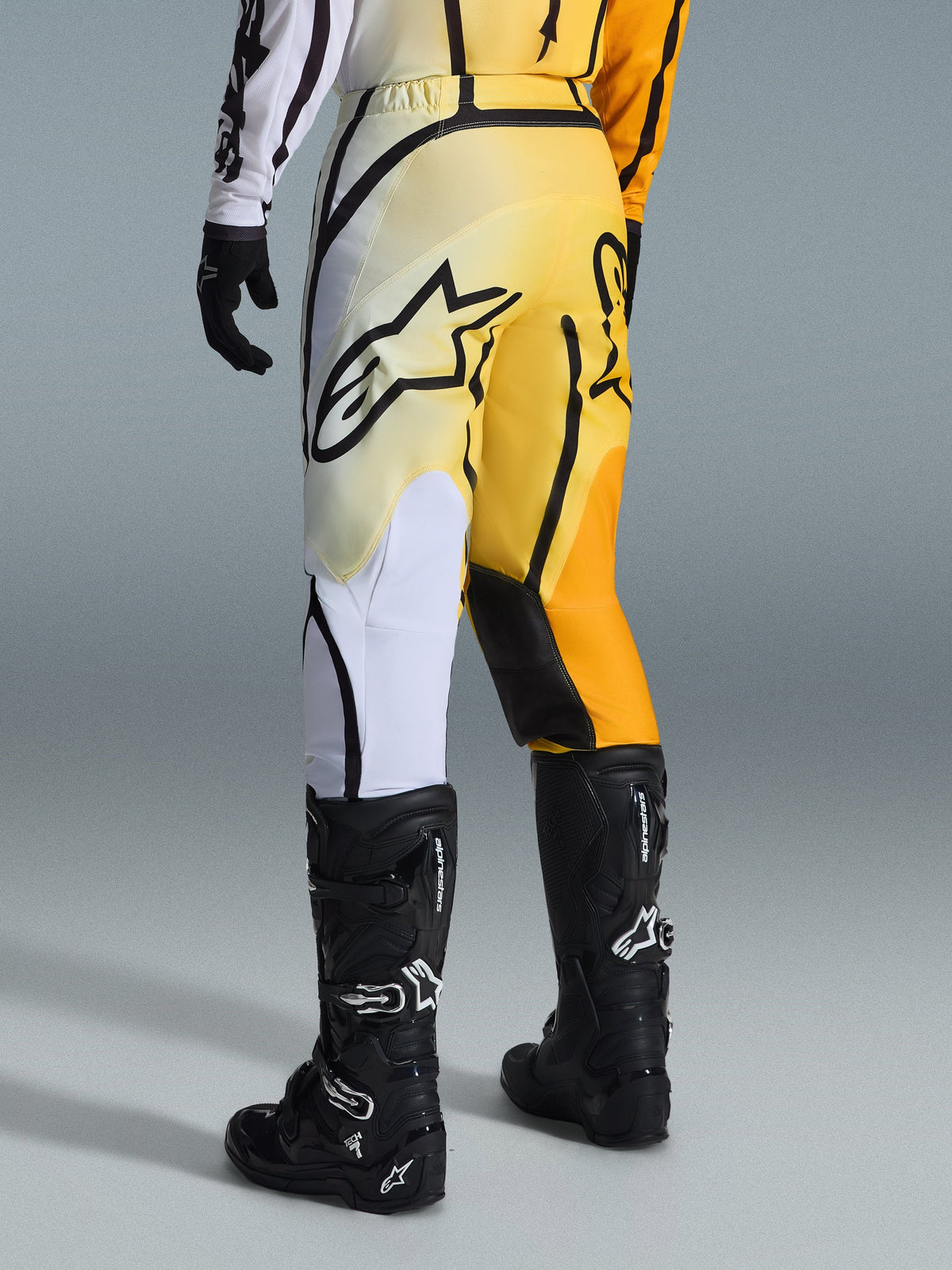 Alpinestars 2026 Fluid Apex Hose, Motocross-Hose, Gelb-schwarzer Farbverlauf mit weißen und orangenen Akzenten, Rückansicht, getragen mit passendem Jersey und schwarzen Tech 7 Stiefeln, markantes schwarzes Alpinestars-Logo auf dem Gesäß, Performance-Ausrüstung für den Geländeeinsatz.