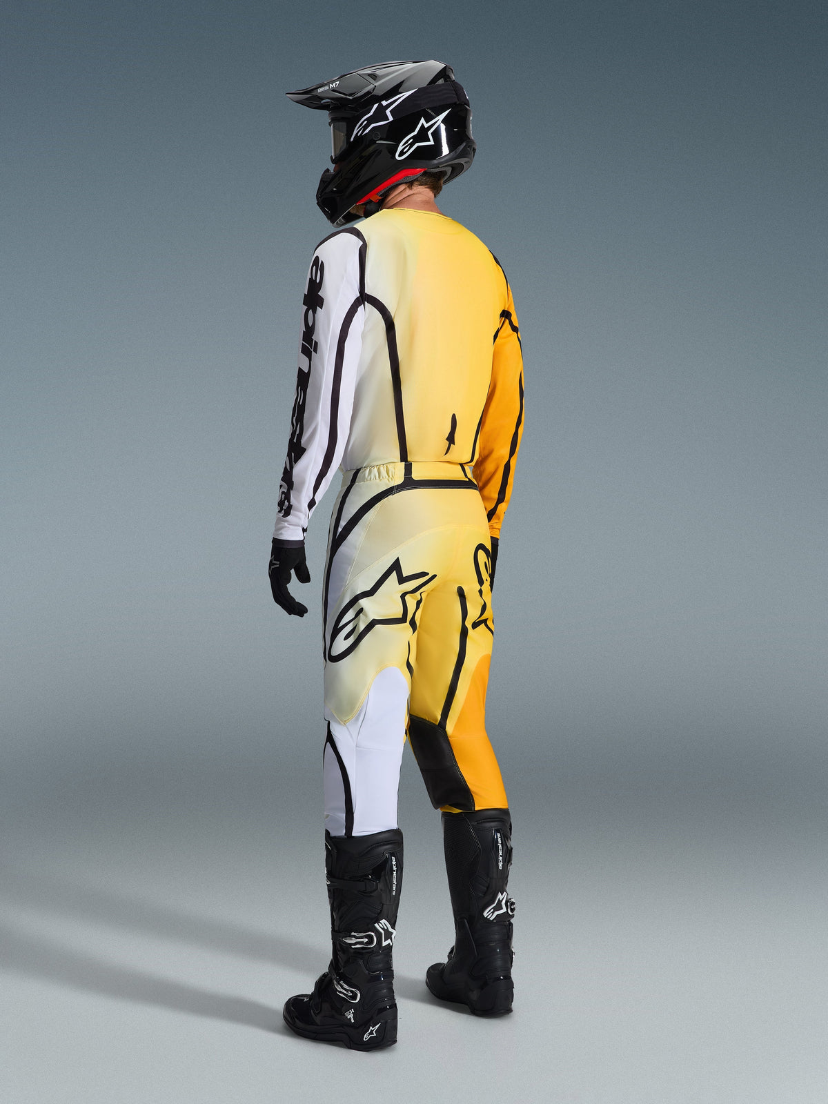 Alpinestars 2026 Fluid Apex Hose, Motocross-Hose, Gelb-Weiß-Farbverlauf mit schwarzen Akzenten, Rückansicht eines Fahrers in kompletter Rennausrüstung einschließlich passendem Jersey, schwarzem Helm und schwarzen Stiefeln