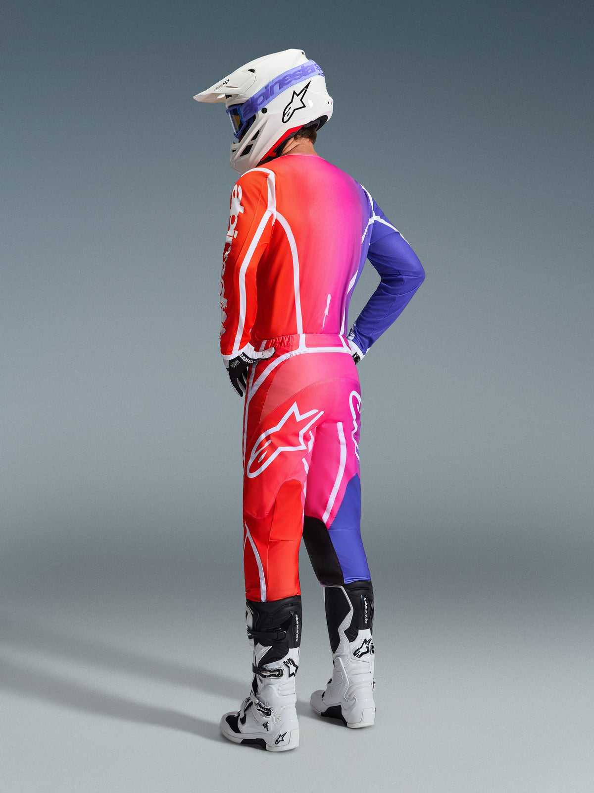 Motocross rider wearing Alpinestars 2026 Fluid Apex gear, Multicolor jersey and pants featuring a red, pink, and purple gradient with white logos, white helmet and white boots, coordinated outfit for off-road racing