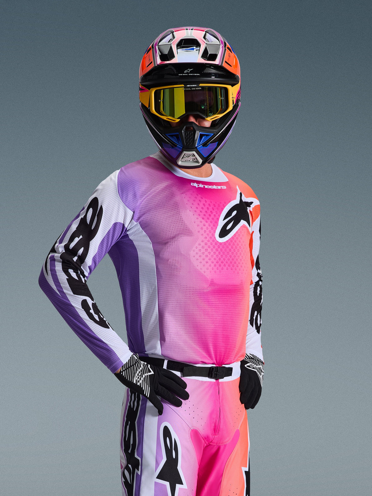 Motocross-Fahrer trägt das Alpinestars 2026 Racer Air Portl Trikot, mehrfarbiger Farbverlauf in Pink, Lila und Orange mit weißen Akzenten, dazu passender bunter Helm und gelbe Brille, schwarze Handschuhe, professionelle Offroad-Rennausrüstung