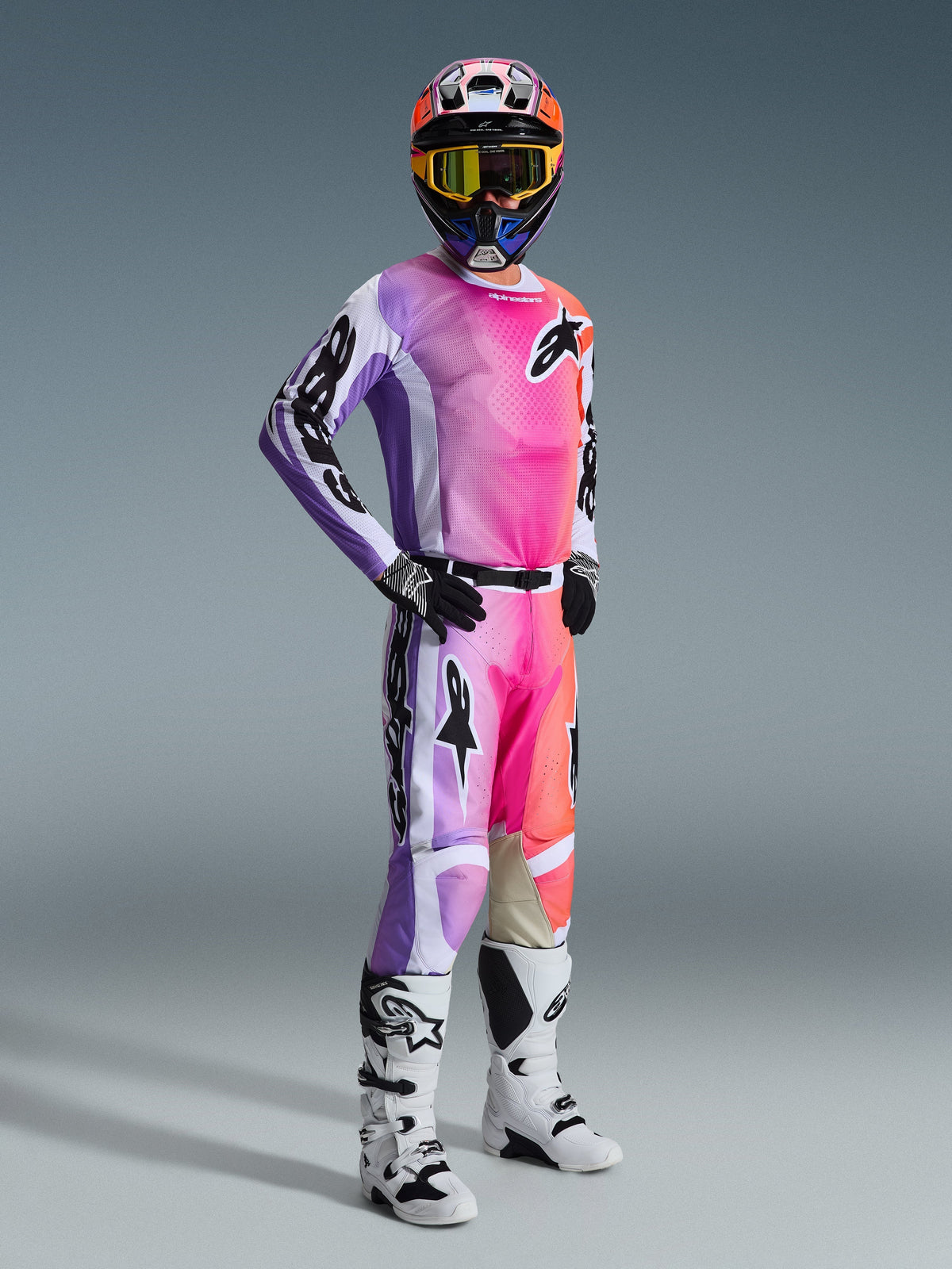 Motocross-Fahrer trägt die Alpinestars 2026 Racer Air Portl Hose in Multicolor mit einem lebendigen Farbverlauf in Pink, Lila und Orange, kombiniert mit einem passenden Trikot, weiß-schwarzen Motocross-Stiefeln und einem abgestimmten Helm mit gelber Brille, komplette Offroad-Rennausrüstung.