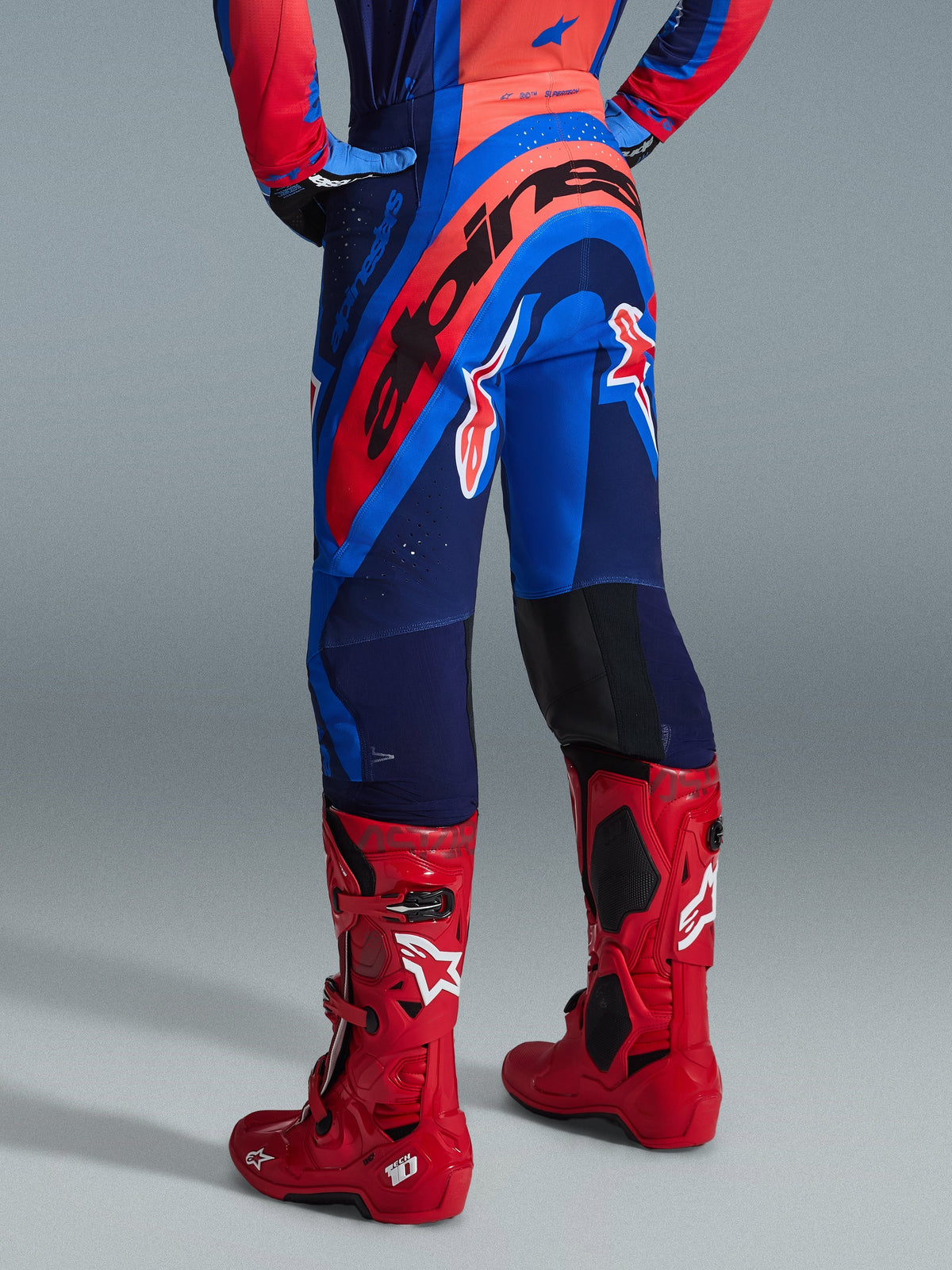 Alpinestars 2026 Supertech Vista Pants, Motocross Pants, Dark Navy Hot Coral Cobalt, back view showing bold red Alpinestars branding across the seat, paired with bright red motocross boots, lightweight stretch construction for professional racing.