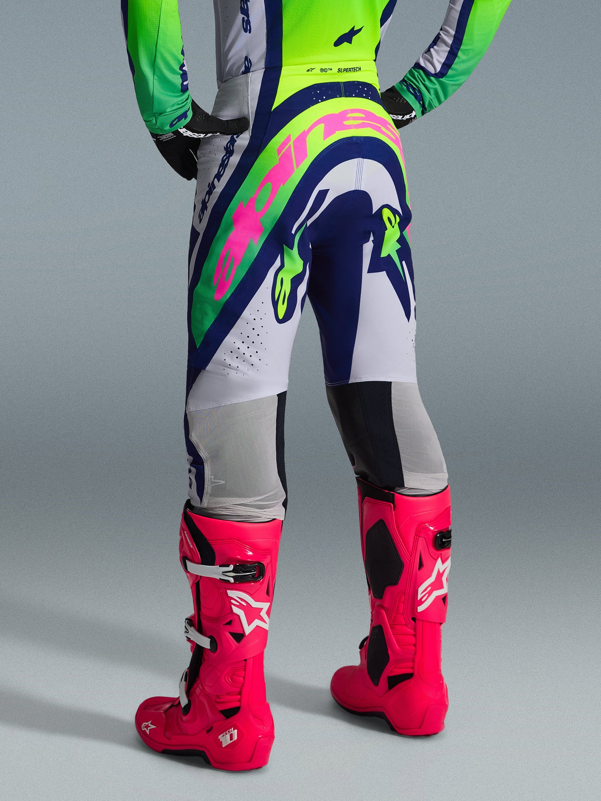 Alpinestars 2026 Supertech Vista Hose, Motocross-Hose, Hellgrau, Gelb Fluo und Blau, Rückansicht mit großem pinkfarbenem Alpinestars-Logo, kombiniert mit passendem Renntrikot und leuchtend pinkfarbenen Tech 10 Stiefeln, professionelle Offroad-Ausrüstung