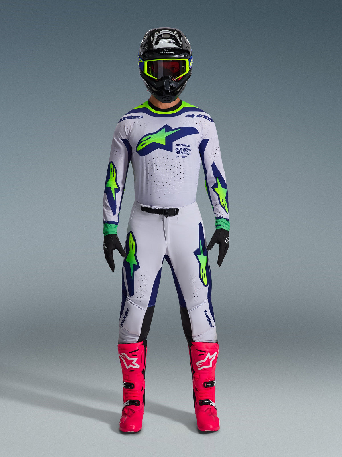 Alpinestars 2026 Supertech Vista Gear Set, Motocross rider wearing complete racing apparel, White jersey and pants with blue and neon green accents, black helmet with neon yellow trim, bright pink boots, professional off-road racing gear ensemble