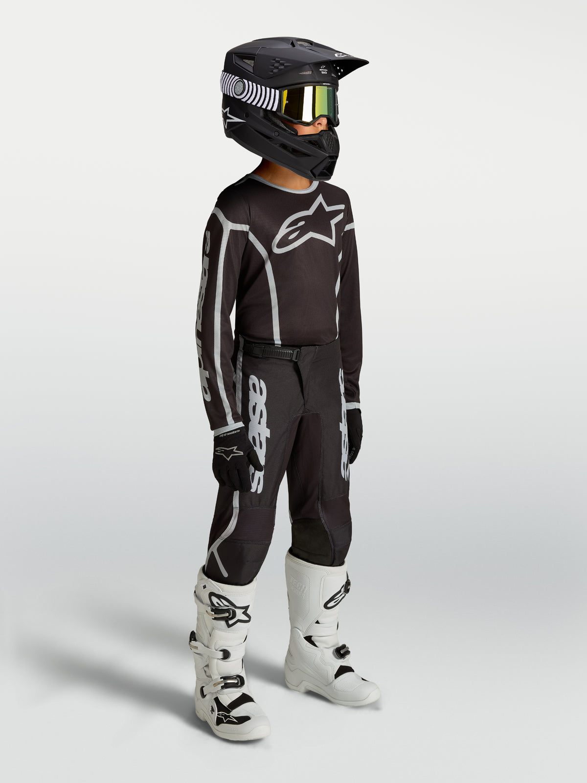 Alpinestars Jugend Fluid Apex V2 Hose, schwarz-silberne Motocross-Ausrüstung am Model mit Trikot, Helm und weißen Stiefeln.