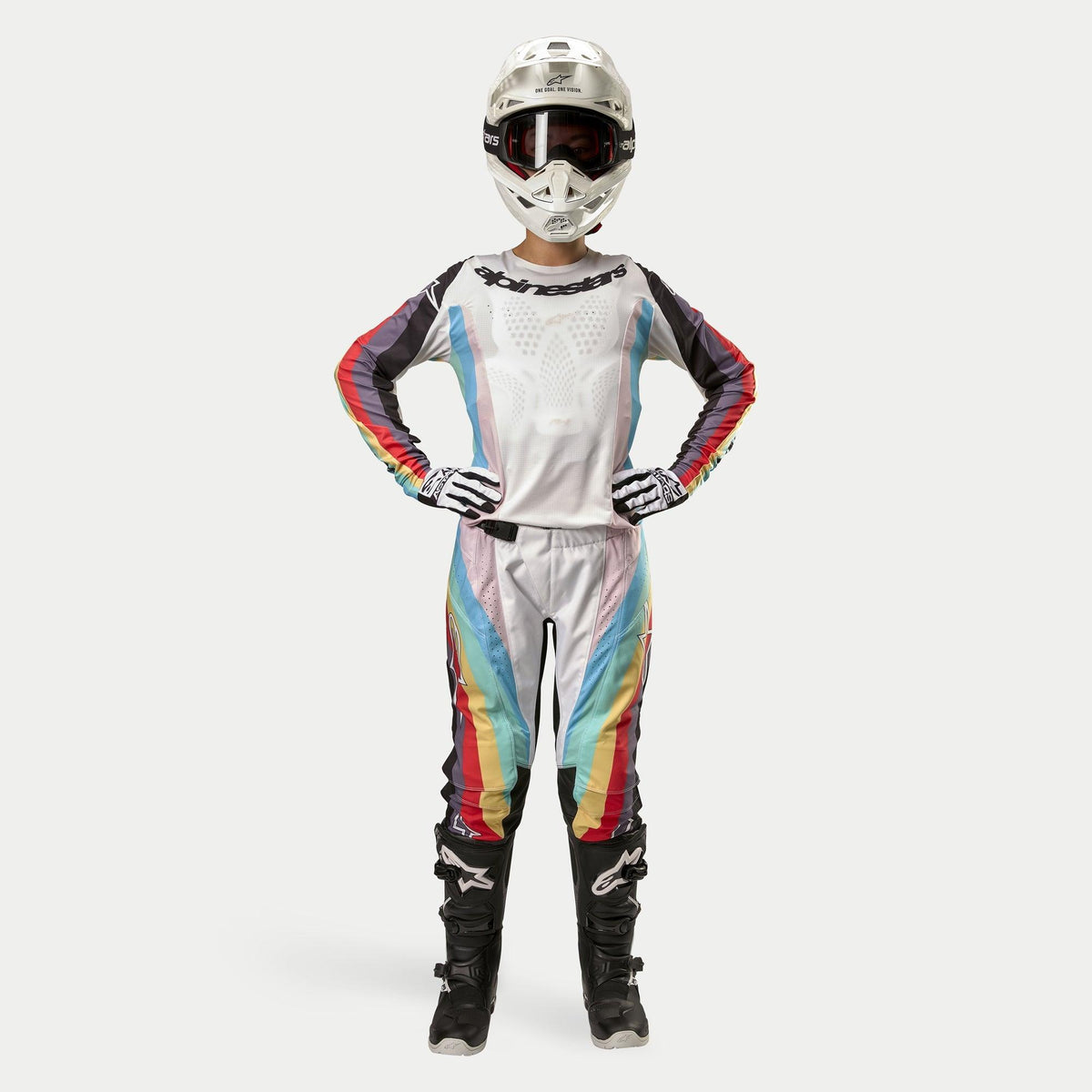 Motocross-Fahrerin in Alpinestars Women Stella 2024 Techstar Hose, Schwarz Multicolor mit weißer Basis sowie roten, gelben und blauen Streifen, passendes Langarmtrikot, weißer Helm und schwarze Stiefel, komplette Rennausrüstung für Offroad-Wettbewerbe