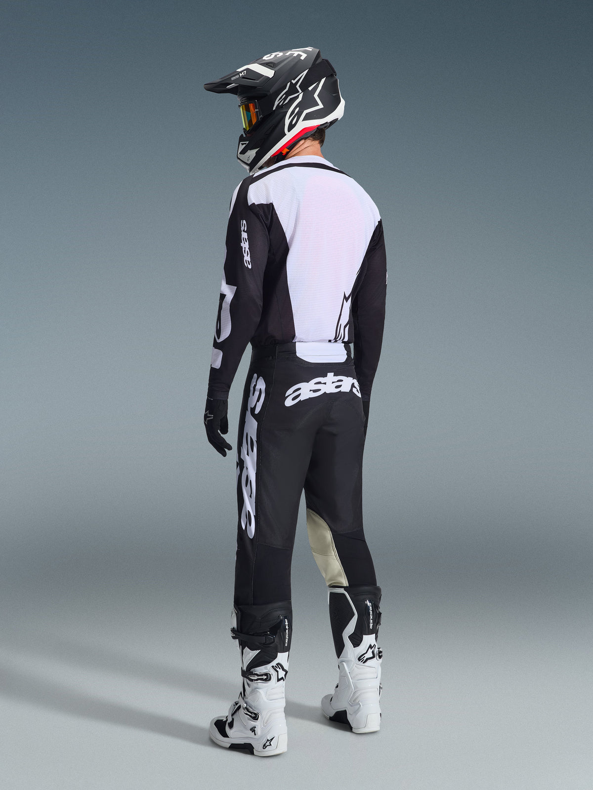 Motocross-Fahrer von hinten in der Alpinestars 2026 Racer Air Riway Ausrüstung in Schwarz und Weiß, mit belüftetem Mesh-Rücken am Trikot und großem weißen Branding, kombiniert mit einem passenden schwarzen Helm und weißen Stiefeln.