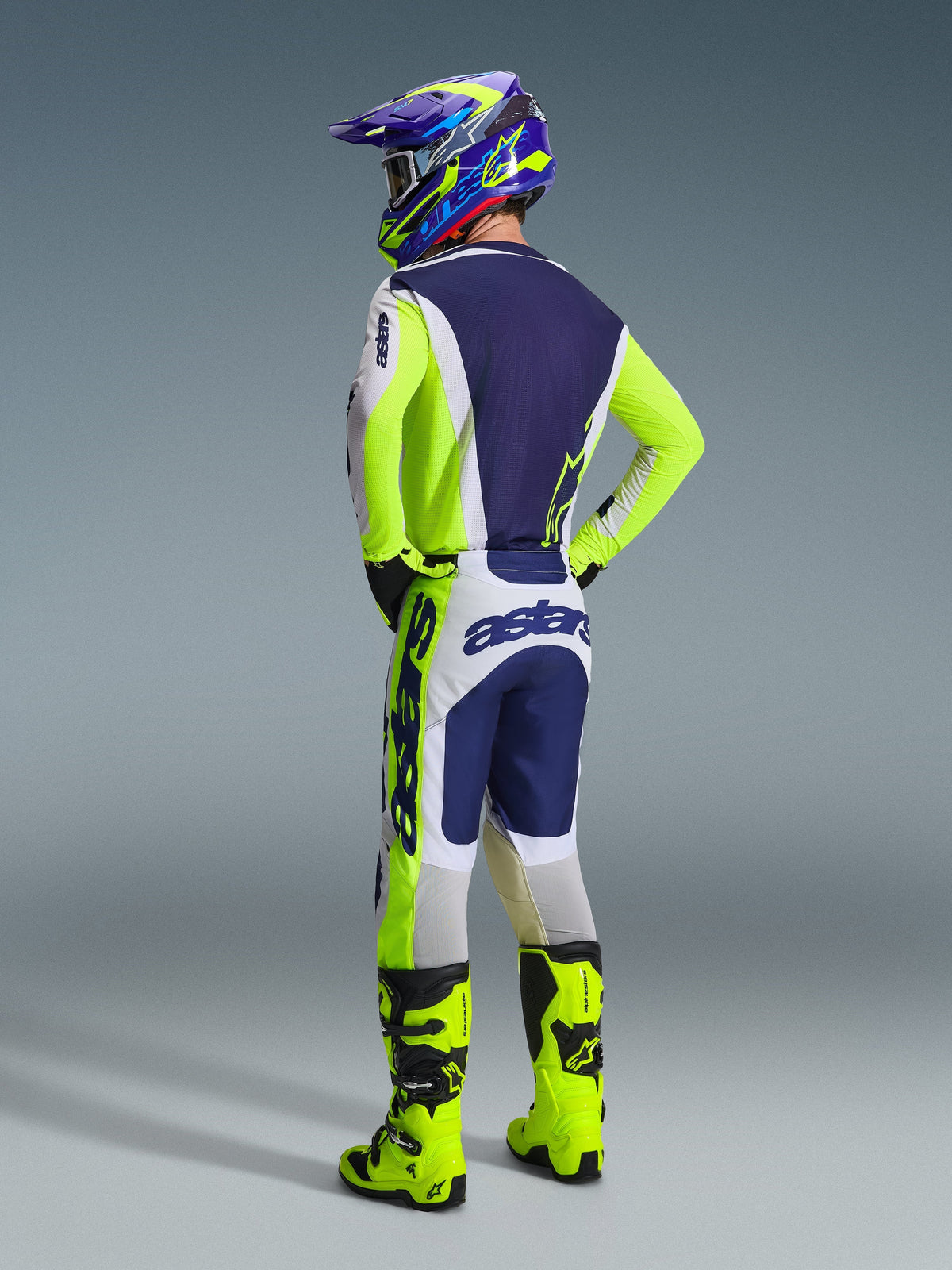 Motocross-Fahrer in der Rückansicht im Alpinestars 2026 Racer Air Riway Gear Set, Trikot und Hose in Hellgrau, Neongelb und Blau, mit „astars“-Branding auf den Ärmeln und am Gesäß, neongelbe Stiefel und ein passender bunter Helm.