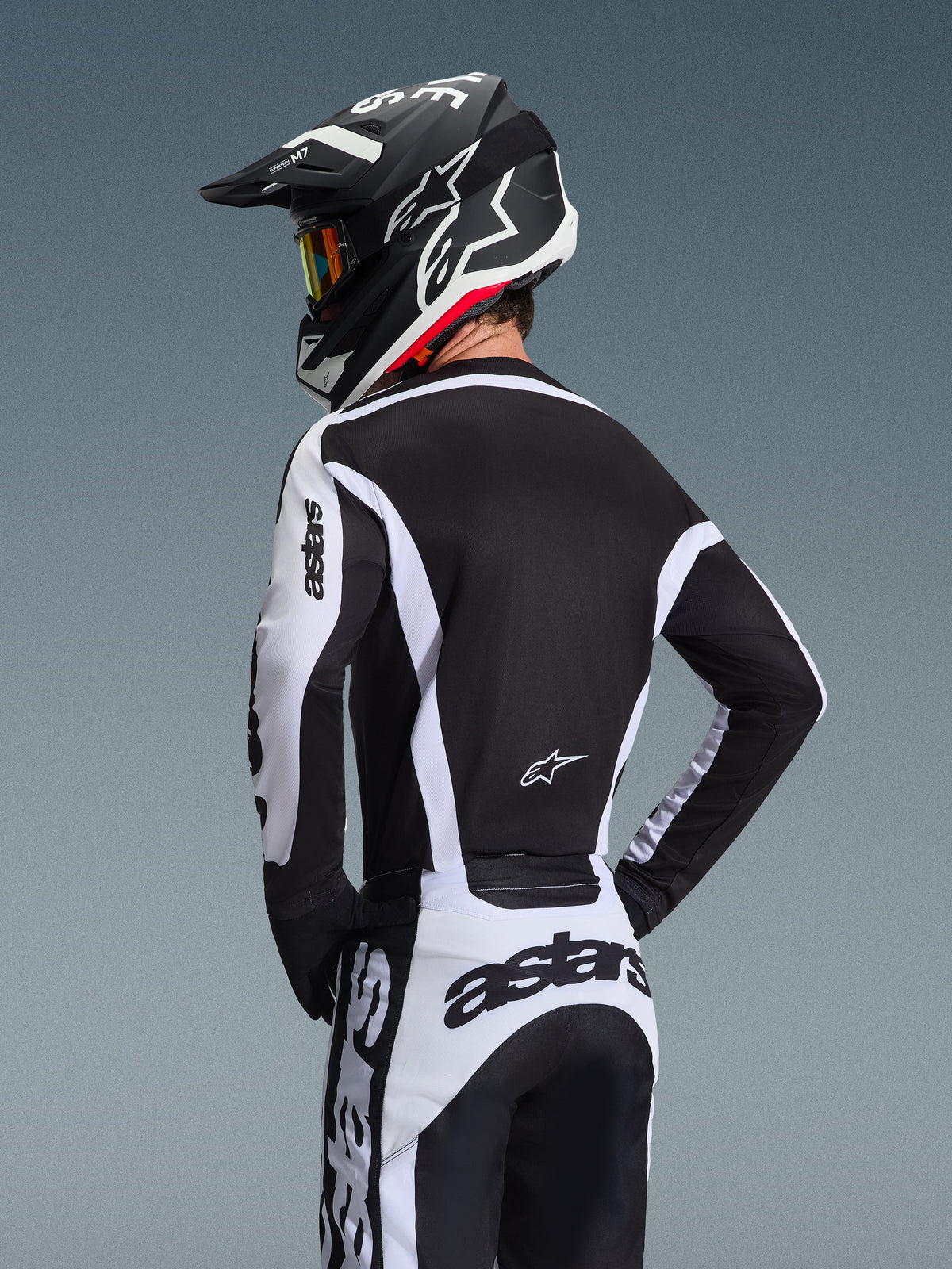 Motocross-Fahrer von hinten im Alpinestars 2026 Racer Riway Trikot in Weiß und Schwarz, mit 'astars'-Branding auf den Ärmeln und dem unteren Rücken, kombiniert mit einer passenden Motocross-Hose und einem schwarzen Helm mit Goggles, abgestimmte Ausrüstung für den Offroad-Rennsport.