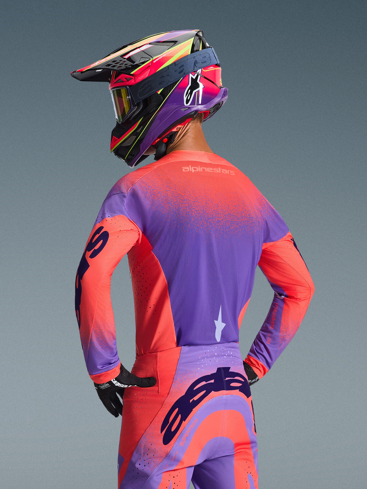 Motocross-Fahrer von hinten im 2026 Alpinestars Supertech Scenz Trikot, Hot Coral und Lila, lebendiges gesprenkeltes Farbverlauf-Design, kombiniert mit passender Motocross-Hose und farbenfrohem Rennhelm, professionelle Ausrüstung für Offroad-Performance