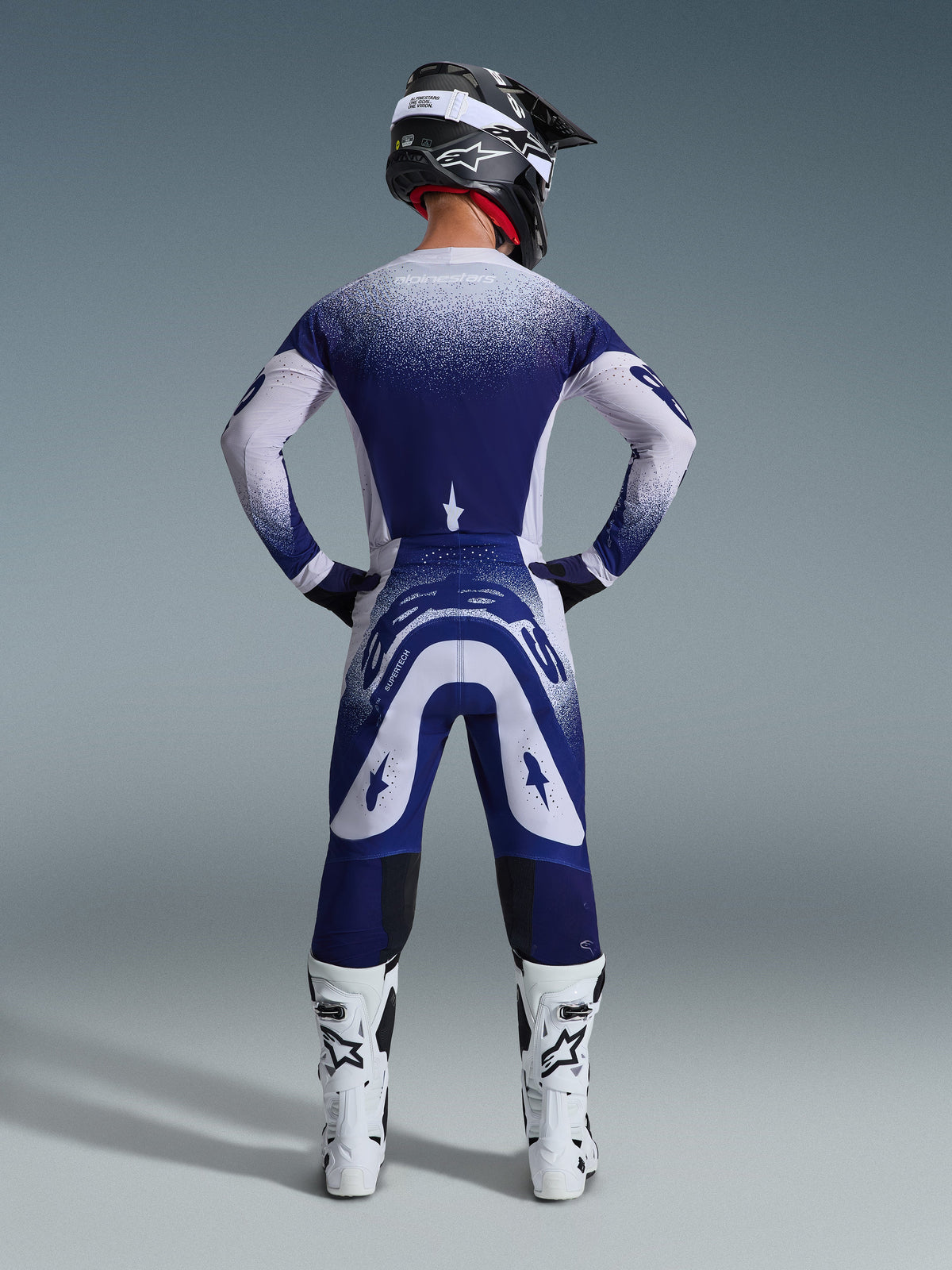 Alpinestars 2026 Supertech Scenz Ausrüstungsset, Rückansicht eines Motocross-Fahrers in blau-weißem Renntrikot und Hose, punktierter Farbverlaufseffekt, weiße Motocross-Stiefel und schwarzer Helm, Profi-Motocross-Ausrüstung