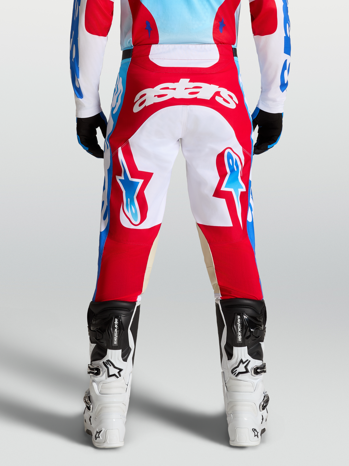 Alpinestars Racer Hose Hunt, Rot und Weiß, Motocross-Hose in der Rückansicht, vorgekrümmte Beinkonstruktion mit hellblauen Akzenten, getragen mit weißen Stiefeln.