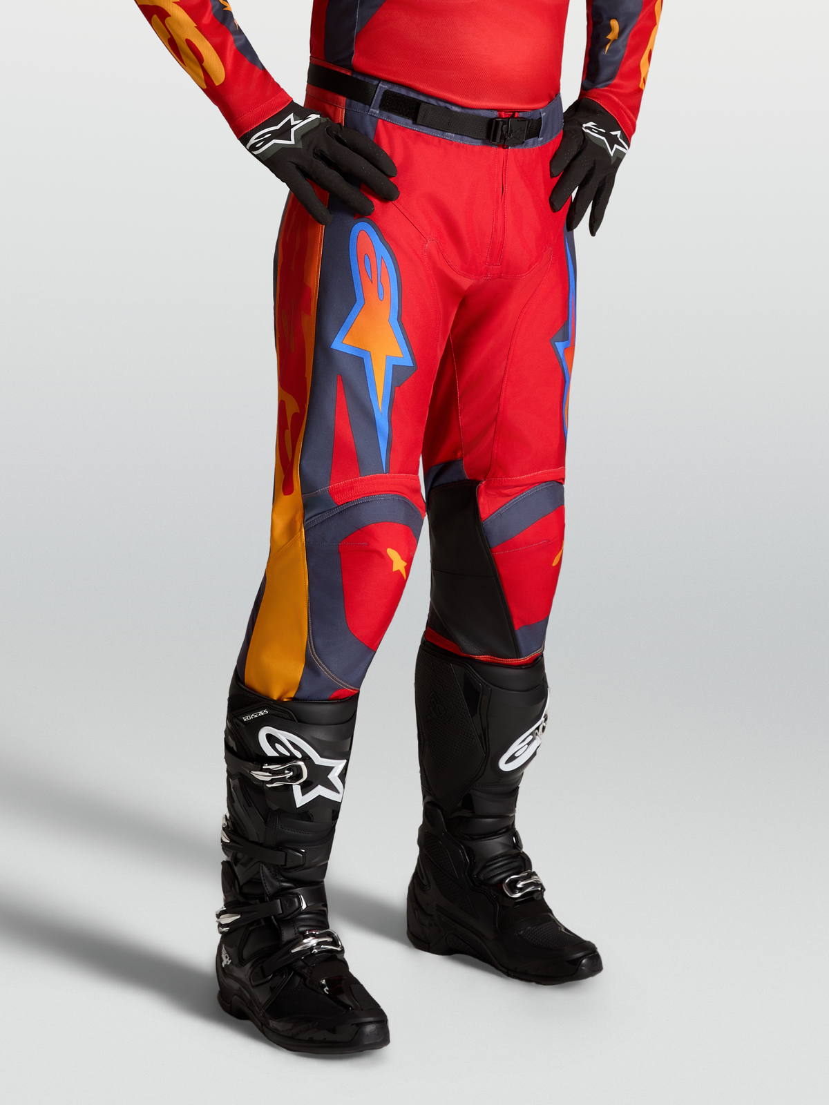 Alpinestars Racer Hunt Hose, Motocross-Hose für Herren, Rot mit grauen und korallenfarbenen Akzenten, am Model mit schwarzen Stiefeln.