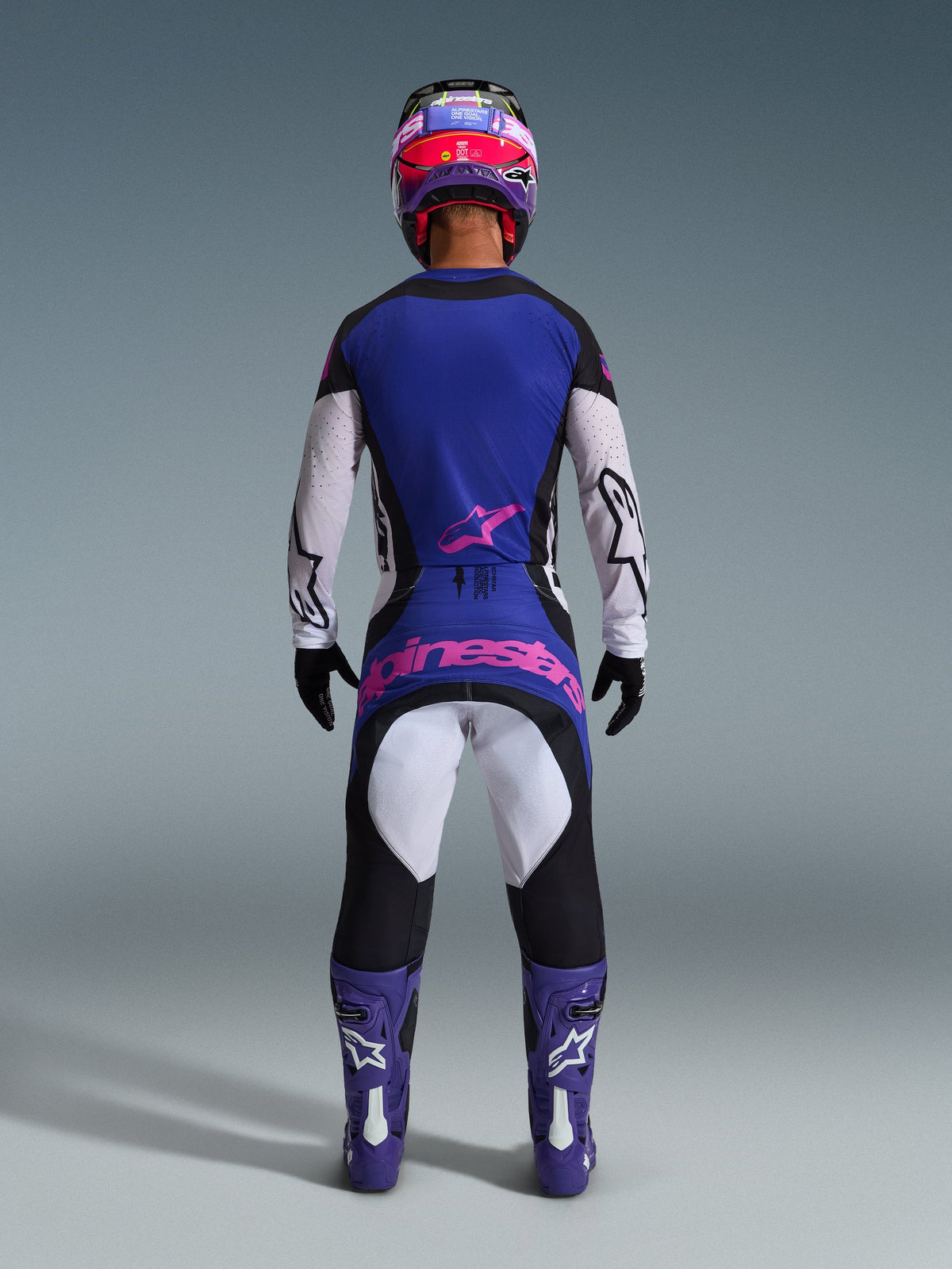 Rear view of a motocross rider wearing the Alpinestars 2026 Techstar Knif Jersey in Black, Light Gray, and Purple, featuring white sleeves and a pink logo on the lower back. Paired with matching motocross pants, purple boots, and a colorful helmet in a studio setting.