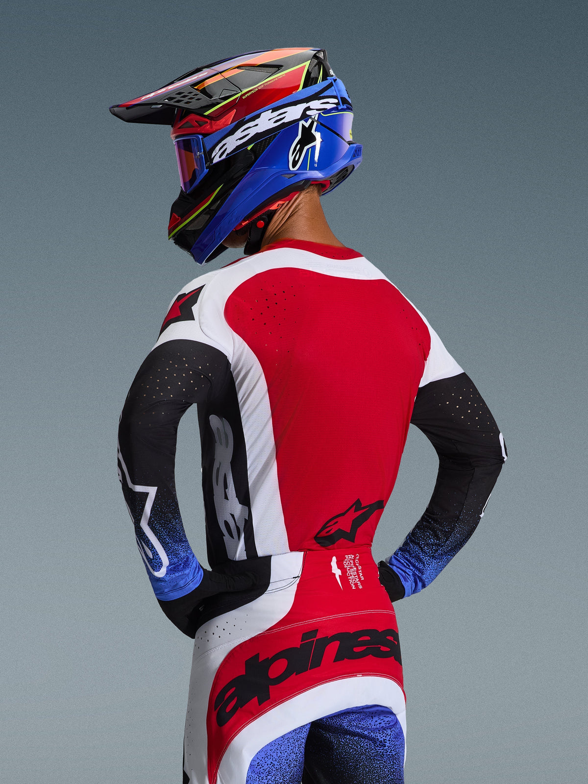 Motocross-Fahrer von hinten gesehen, trägt das 2026 Techstar Knif Jersey in Rot, Weiß und Schwarz, mit lasergeschnittenen Perforationen am Rücken und markantem Branding, kombiniert mit einer passenden Hose mit blauen Akzenten und einem blau-schwarzen Alpinestars-Helm, professionelle Offroad-Rennsport-Ästhetik