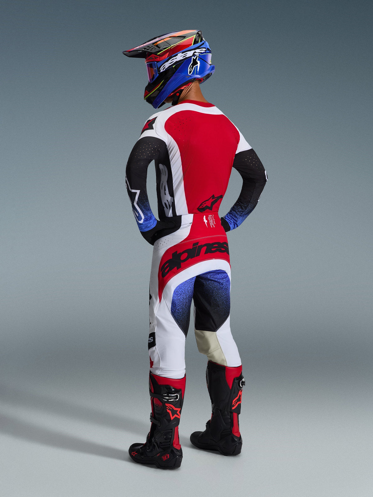Motocross rider seen from the back wearing complete off-road gear, 2026 Techstar Knif Jersey in Red White and Black with blue sleeve details, matching technical pants, blue racing helmet, and black and red boots, professional motocross racing kit.