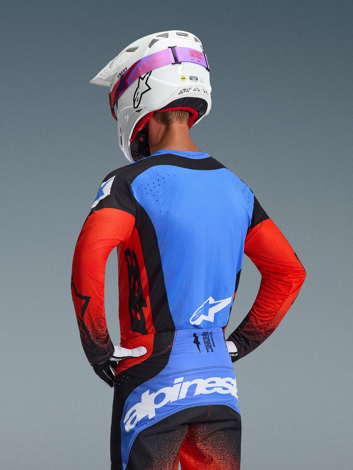 Motocross-Fahrer in der Rückansicht im 2026 Techstar Knif Gear Set, blaues Jersey mit roten Ärmeln und schwarzen Akzenten, schwarz-blaue Hose mit weißem Alpinestars-Branding, weißer Helm mit pinkem Brillenband, lasergeschnittene Perforationen zur Belüftung