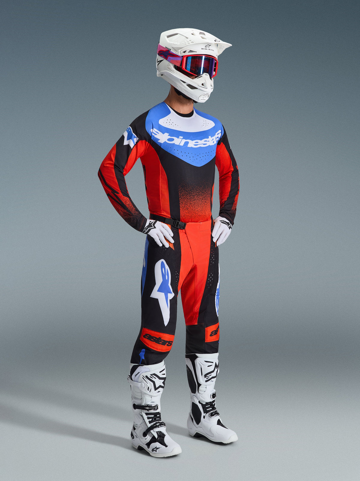 Motocross-Fahrer im Alpinestars 2026 Techstar Knif Gear Set, Trikot mit blauem Brustbereich, weißem Logo und roten Ärmeln mit schwarzen Farbverläufen, passend dazu schwarz-rote Hose mit blauen Stern-Akzenten, weißer Helm mit rot-blauer Schutzbrille, weiß-schwarze Stiefel und weiße Handschuhe.