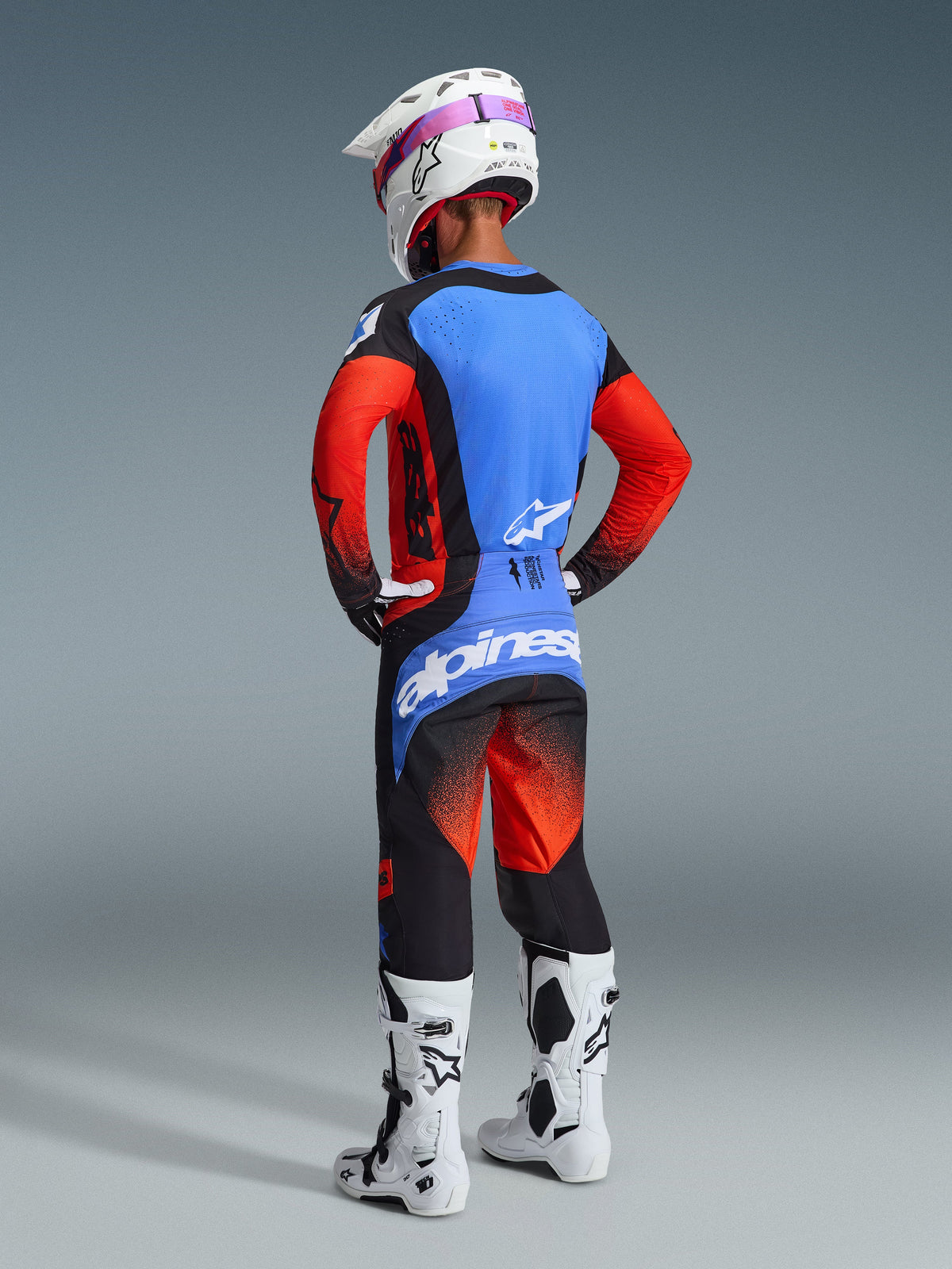 Rückansicht eines Motocross-Fahrers im Alpinestars 2026 Techstar Knif Gear Set, bestehend aus einem blau-roten Jersey mit schwarzen Akzenten, passender schwarz-roter Hose mit großem blauem Logo auf der Sitzfläche, weißem Helm und weißen Motocross-Stiefeln, professionelle Offroad-Rennausrüstung